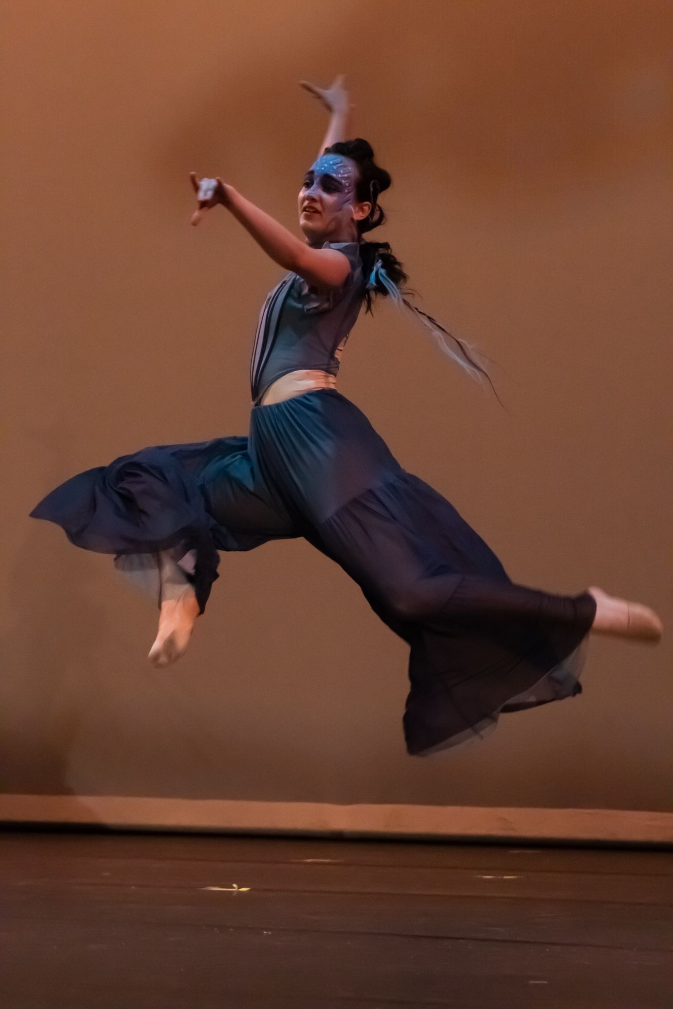 A masked dancer leaps dramatically mid-air in a flowing dark skirt and teal costume during a stage performance.