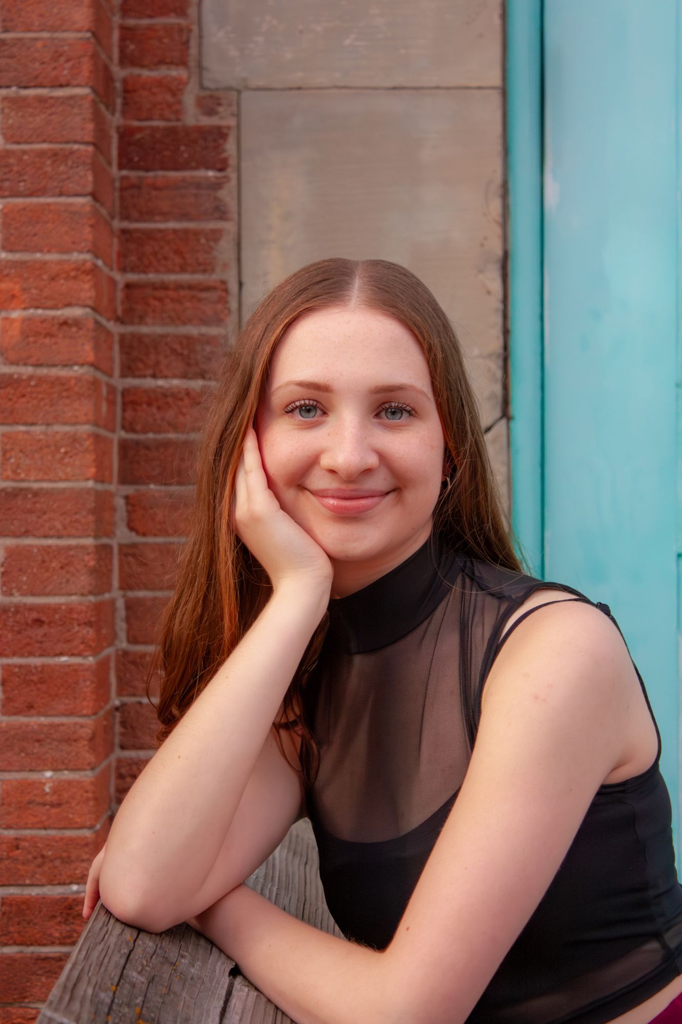 Young woman with auburn hair in black sheer top smiles while leaning against brick wall.