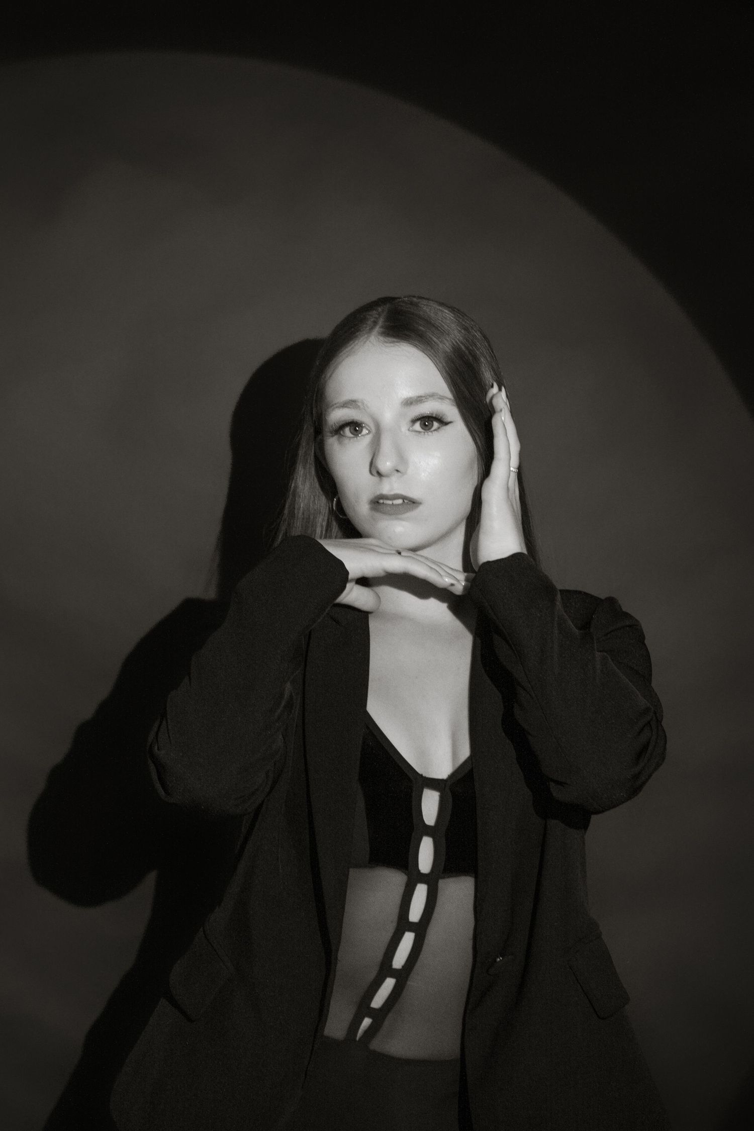 Black and white portrait of young woman in black blazer posing with hands near face under circular light.