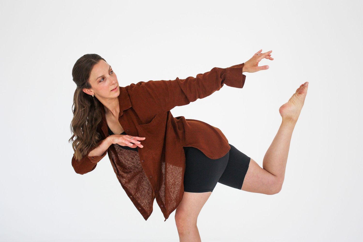 A female dancer in a brown jacket and black shorts performs a dynamic kick pose against a white background. Branding photography Tampa, FL.