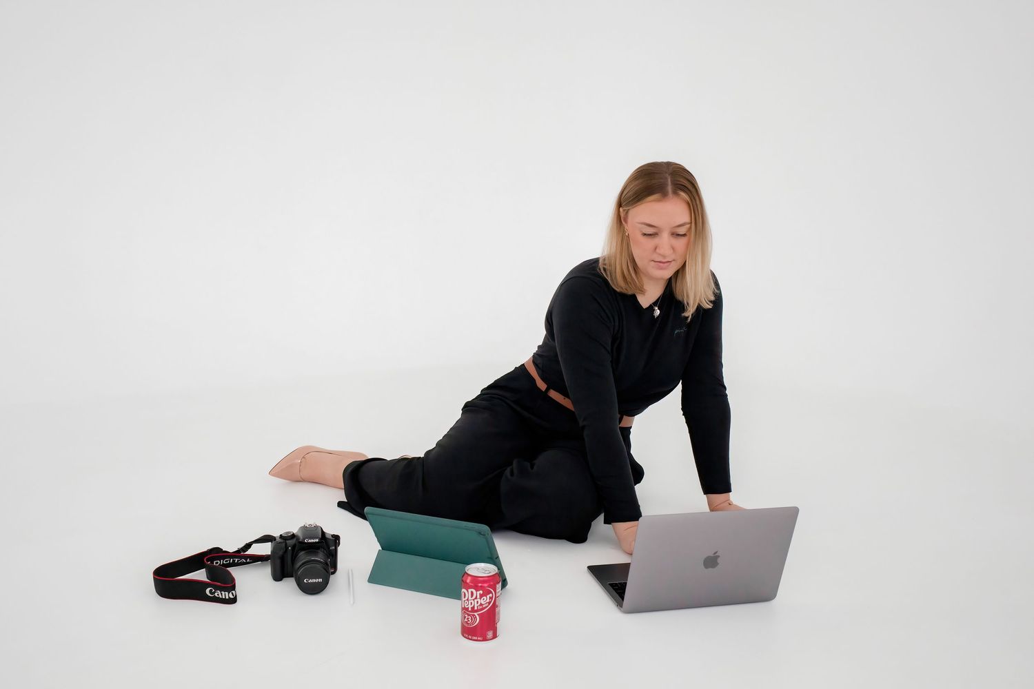 Woman in black outfit sitting on floor using MacBook laptop with Canon camera and drink nearby.