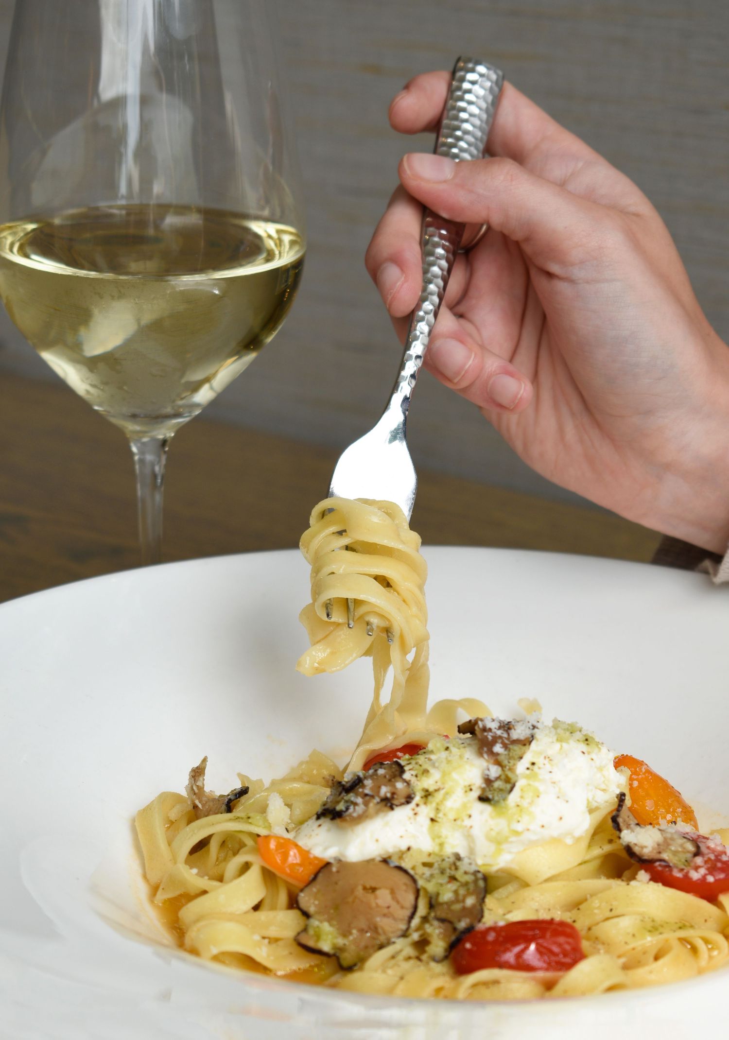 Hand twirling fettuccine pasta with burrata, cherry tomatoes, and mushrooms beside a glass of white wine.