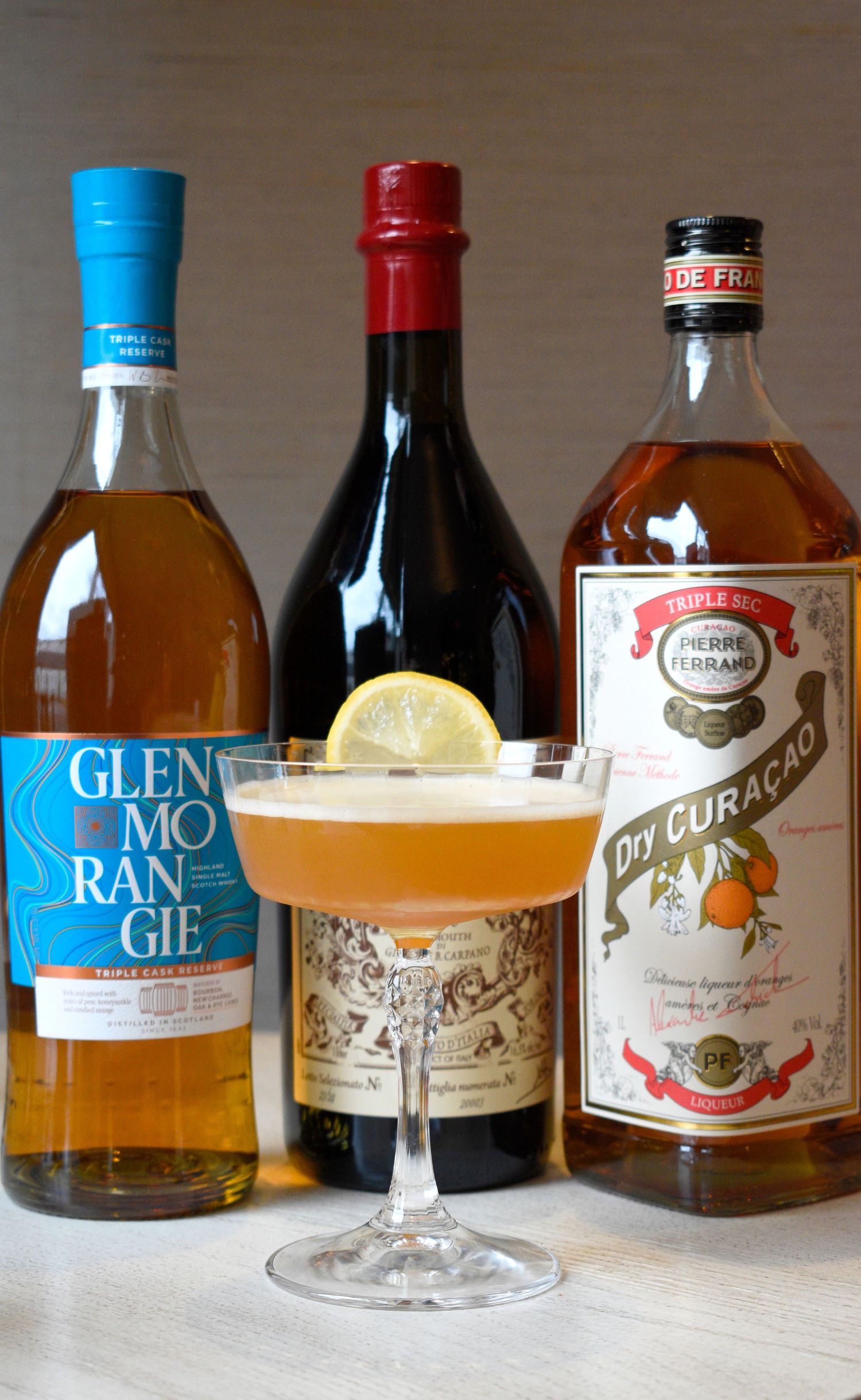 Amber sour cocktail with lemon garnish surrounded by Glenmorangie whisky, red wine, and Pierre Ferrand Dry Curacao bottles.