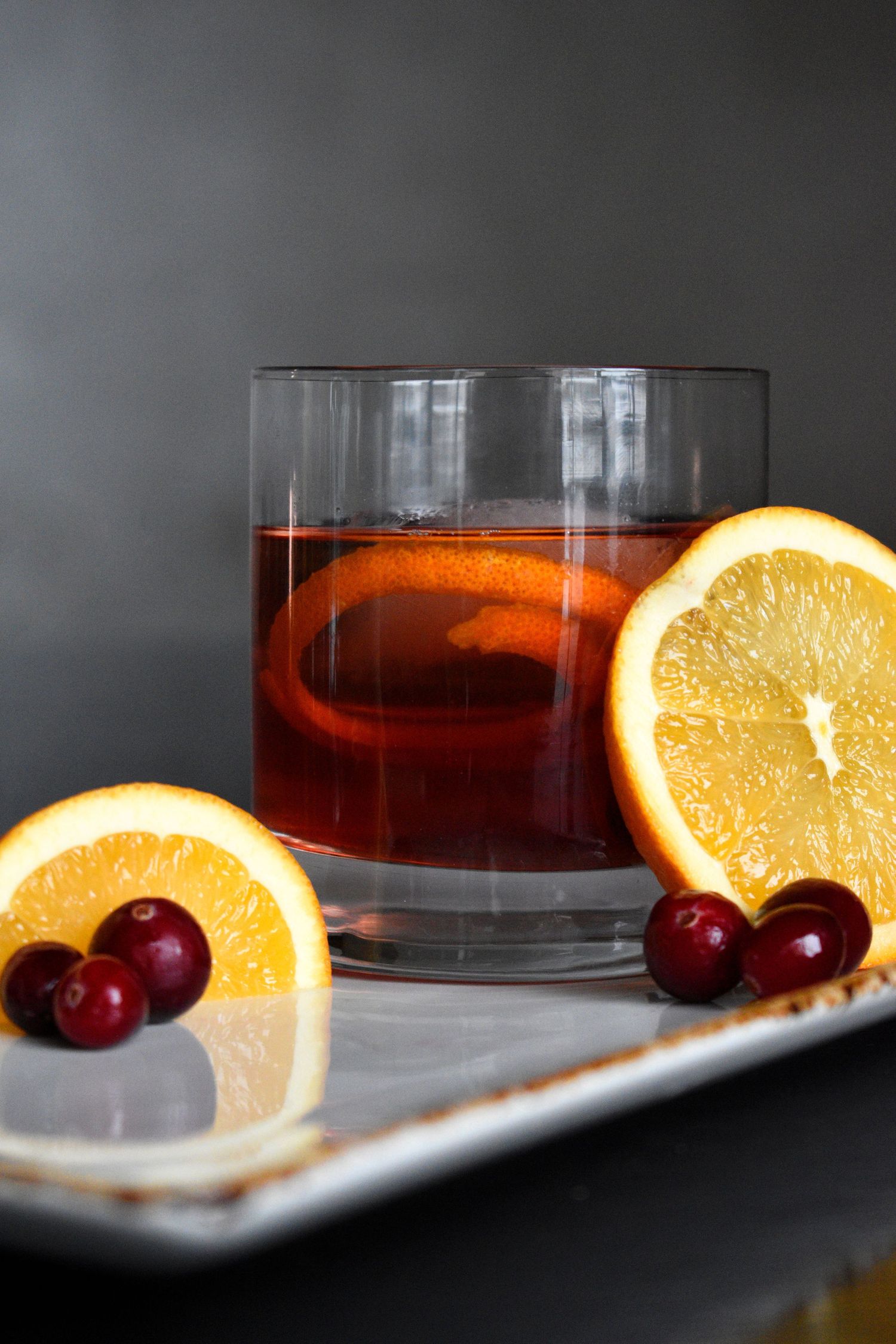 A dark red cocktail on a white tray garnished with fresh cranberries and orange slices.