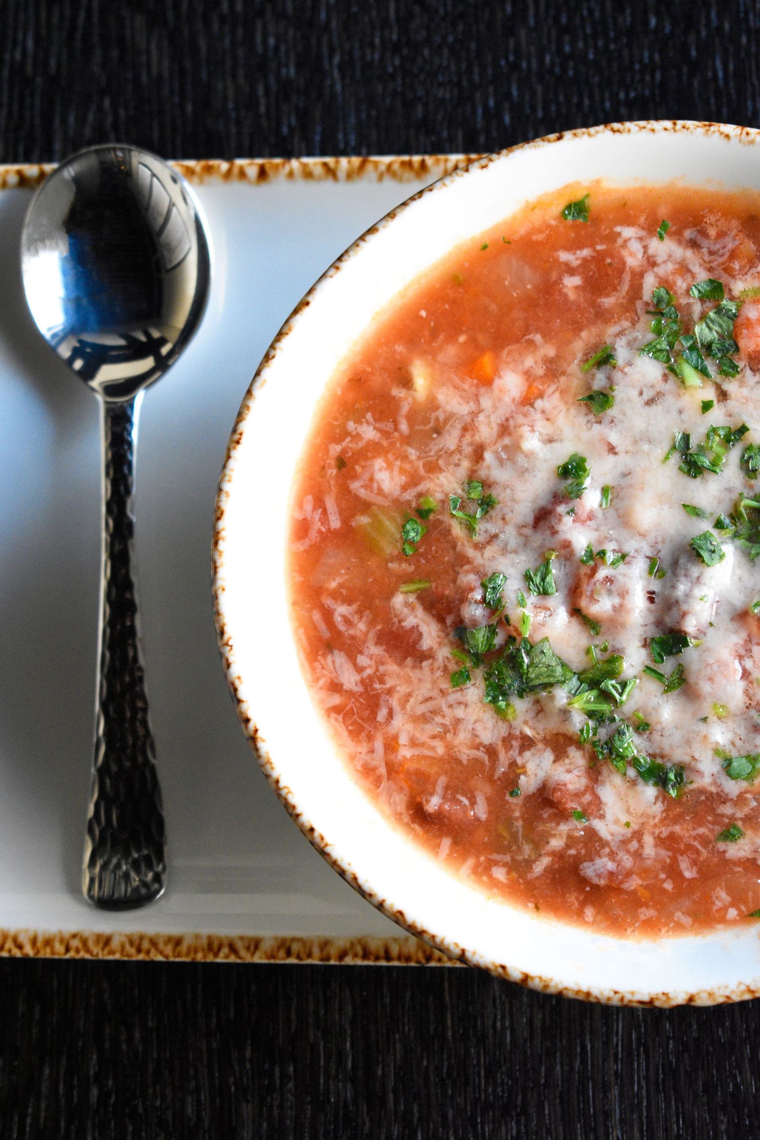 A bowl of tomato-based soup with rice, garnished with parsley, served with a spoon on a white plate.