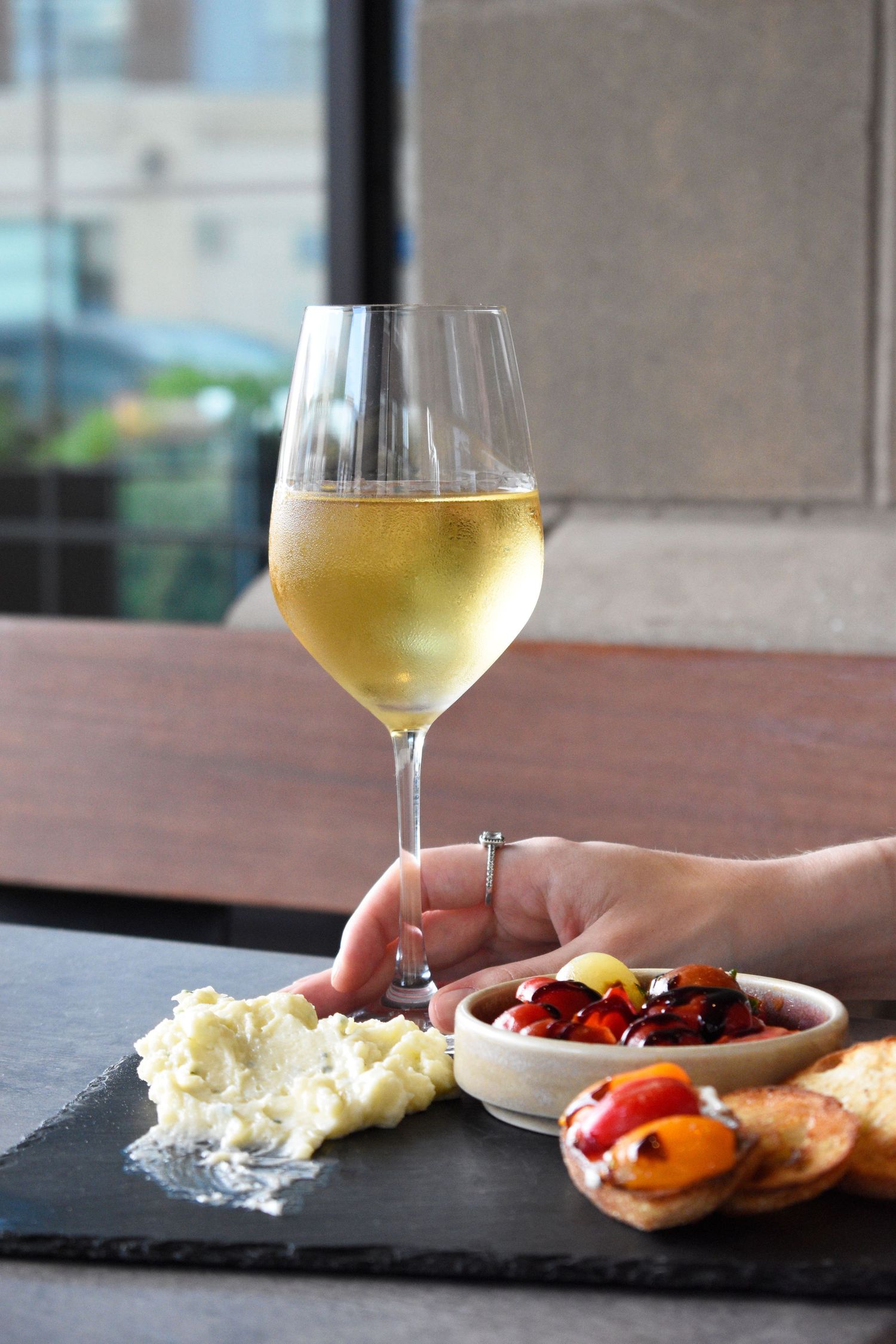 Hand holding white wine glass on slate board with whipped cheese, marinated peppers, and toasted bread. Food & beverage photography in Tampa