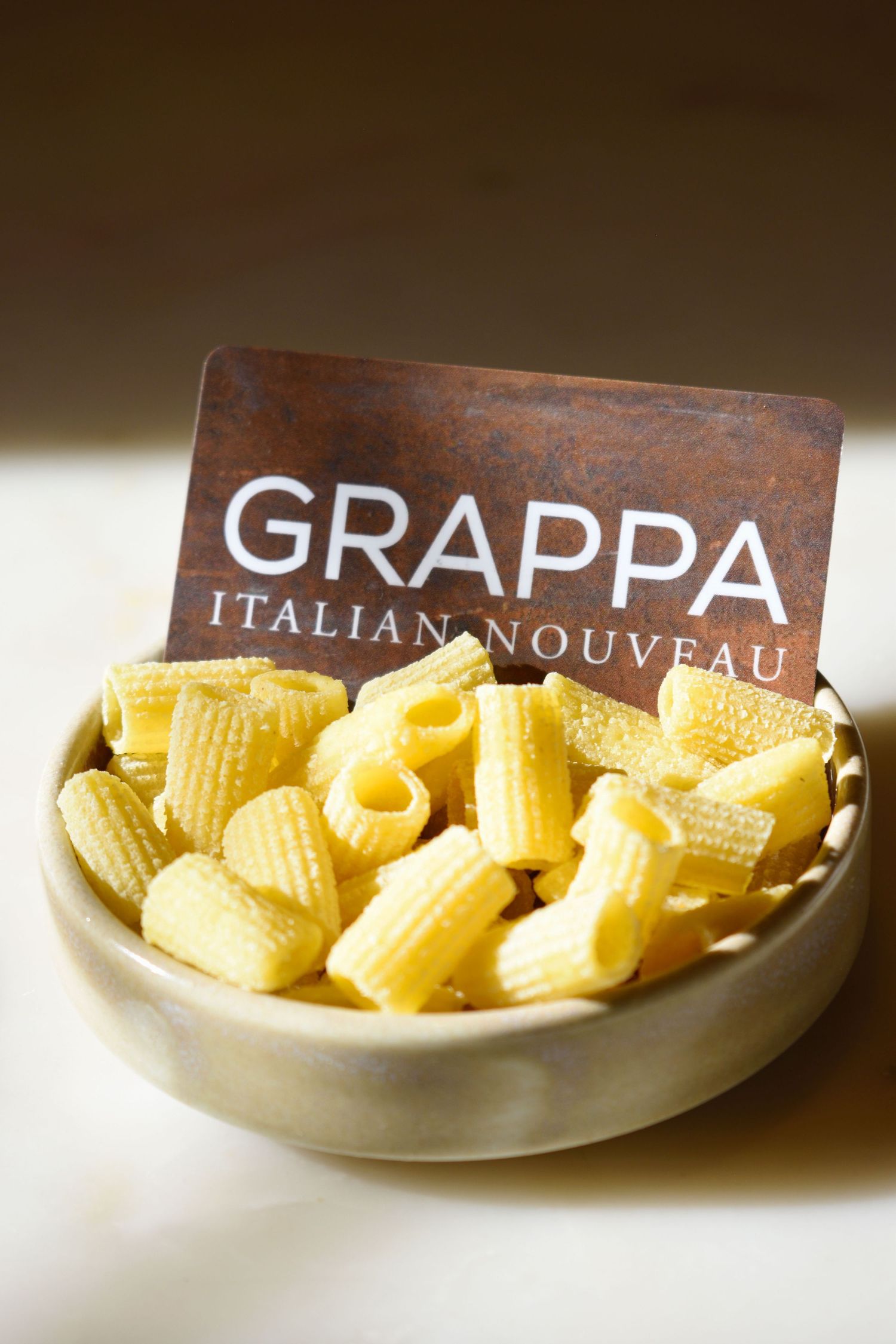 Grappa Italian Nouveau gift card displayed in bowl of uncooked rigatoni pasta on white surface.