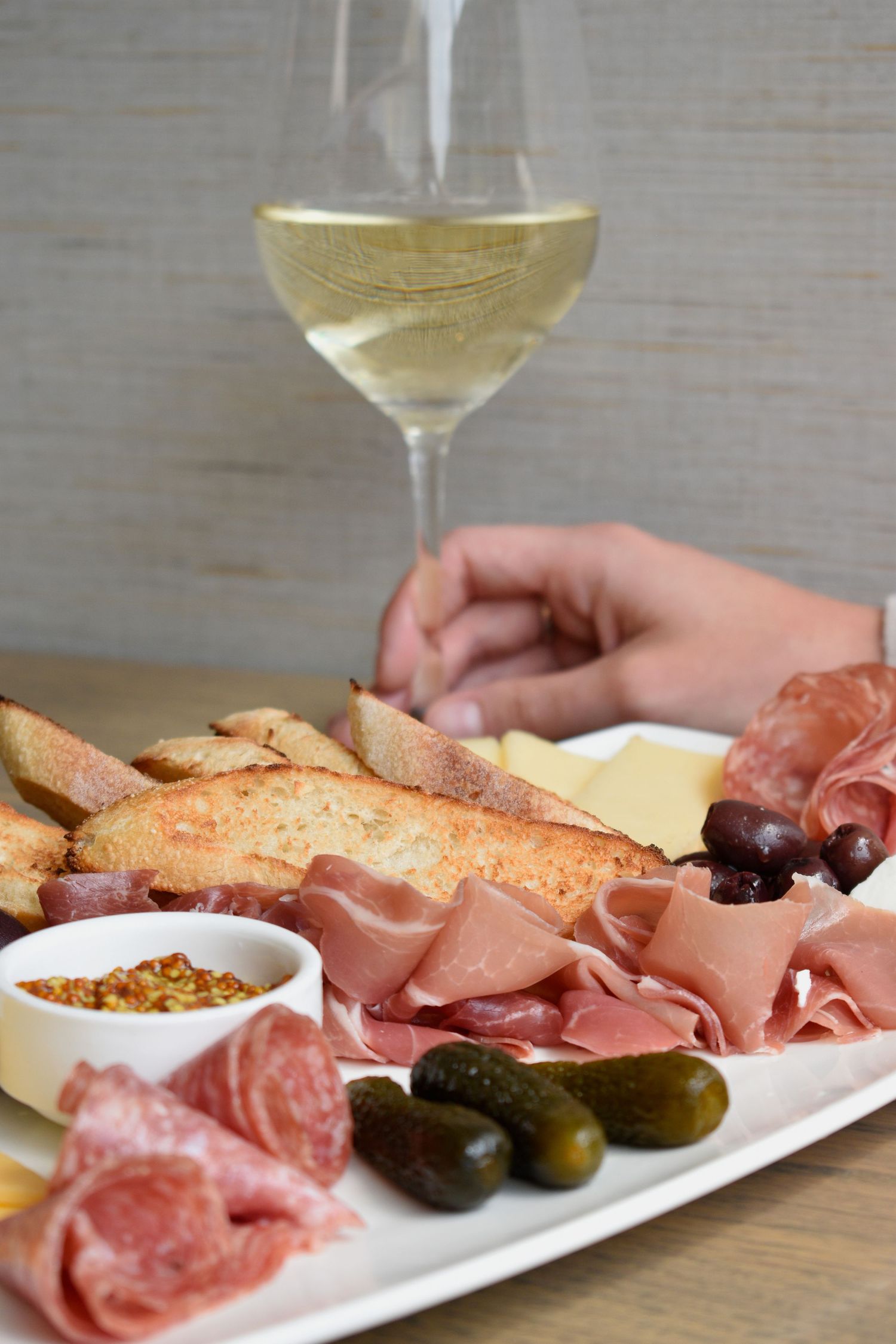 Charcuterie board with prosciutto, salami, cheese, olives, pickles, and baguette slices paired with white wine.