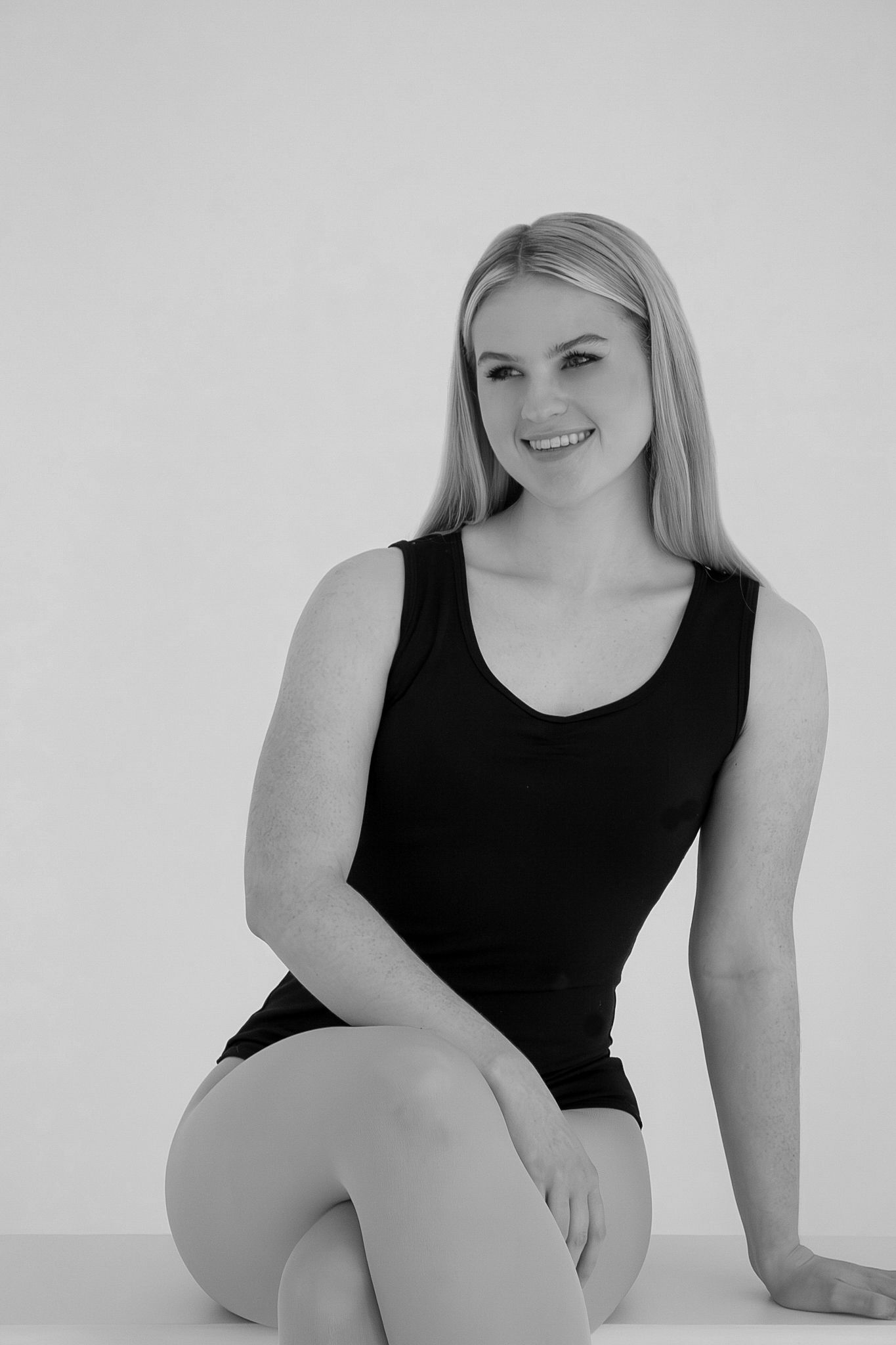 Smiling blonde woman in black bodysuit sitting casually in black and white photo.