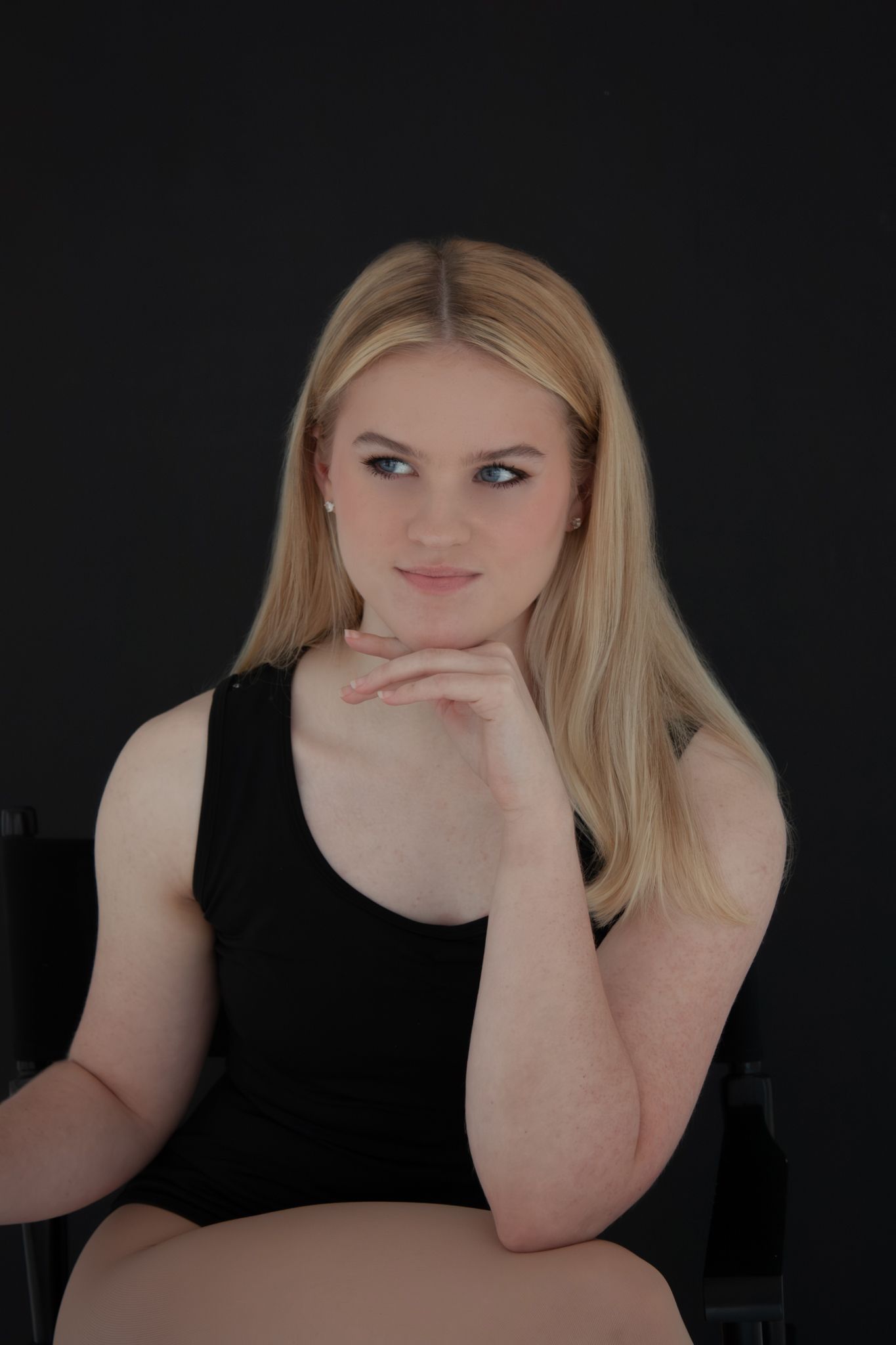 Young blonde woman in black sleeveless top sits confidently with hand on chin against dark background.