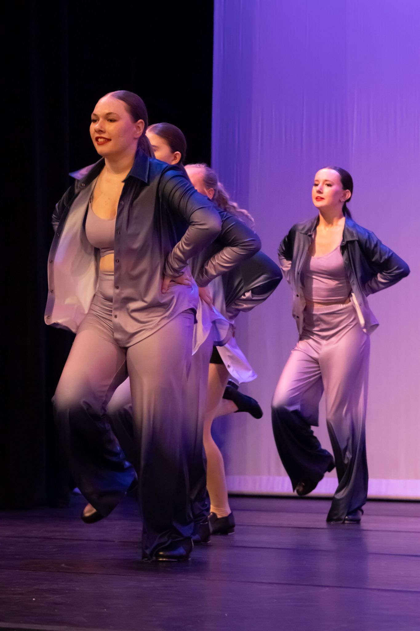Teen dancers in gray outfits and black jackets perform a jazz dance routine on a dark stage.