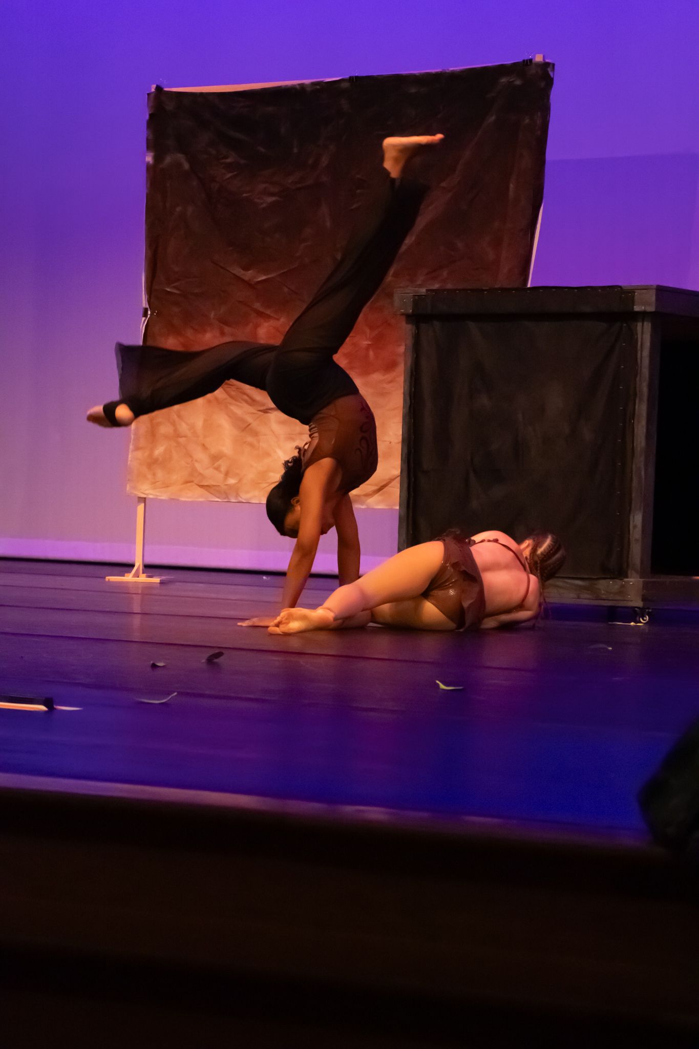 Two dancers perform on a purple-lit stage, one doing a handstand and another lying on the floor.