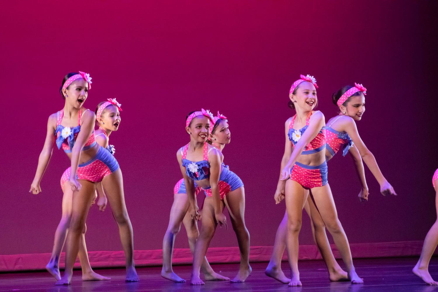A group of young girls in pink and blue dance costumes perform an upbeat routine on a pink-lit stage.