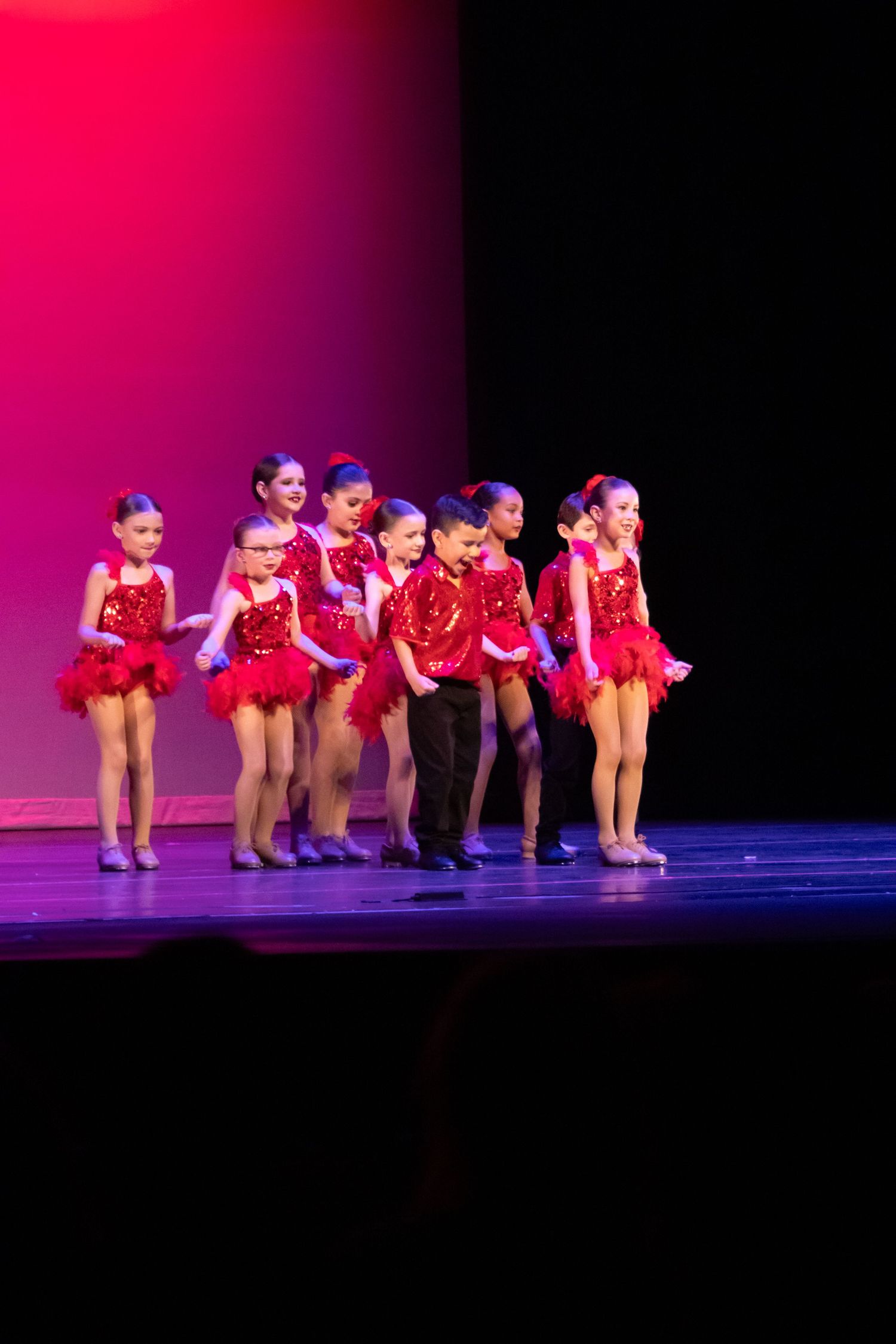 Young dancers in red sequin costumes perform on stage under pink spotlight at a dance recital.