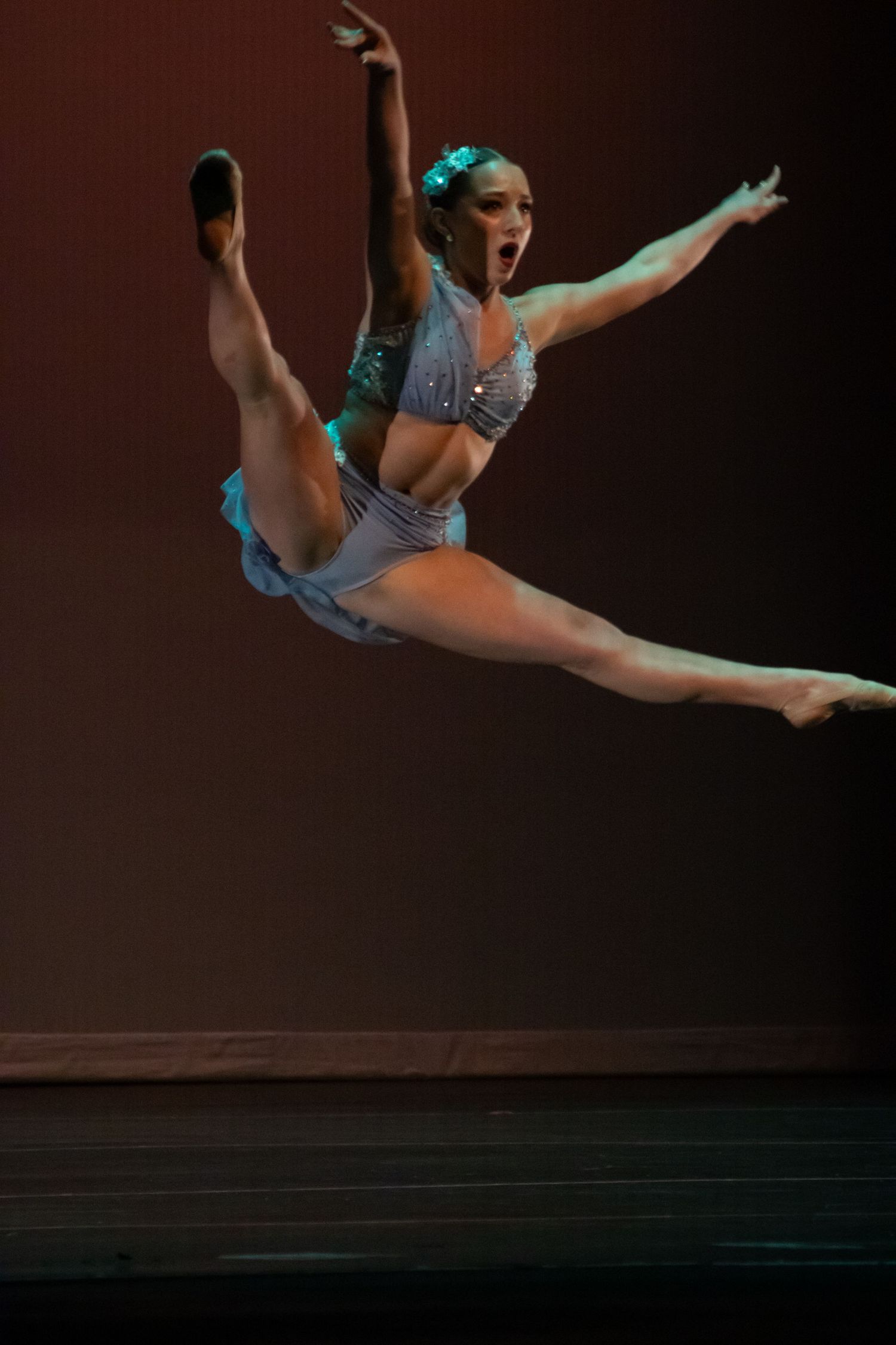 Dancer in silver and teal costume executes spectacular leap split under warm stage lighting.