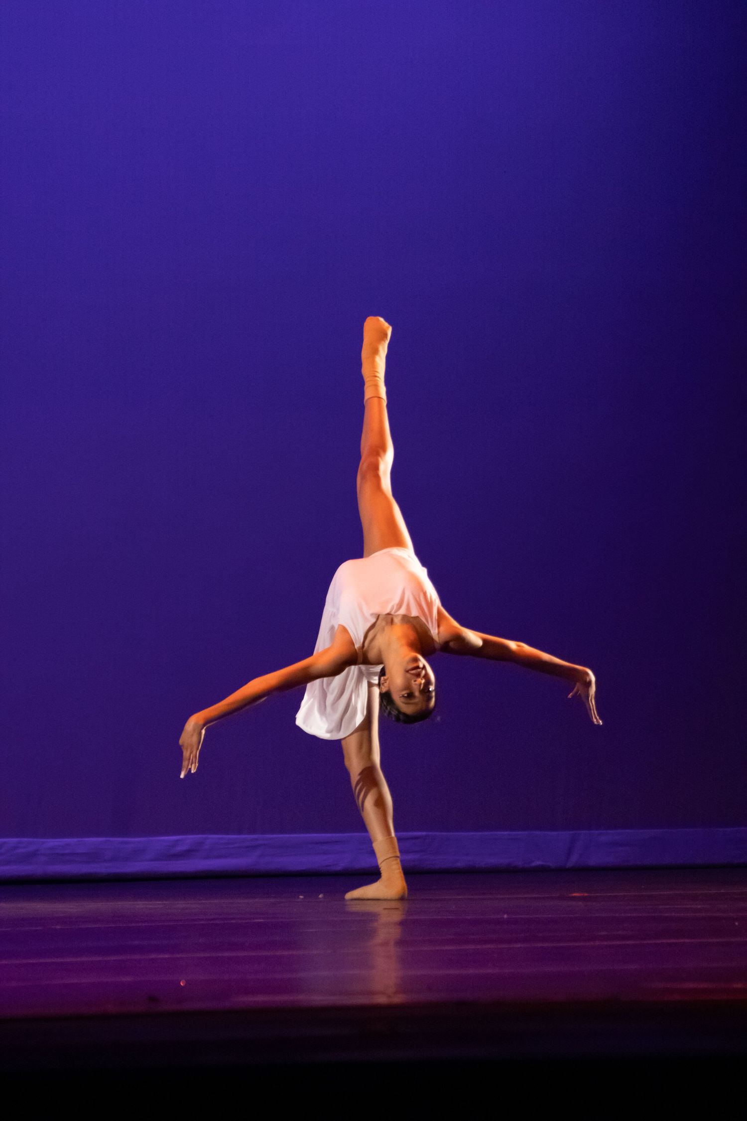 A dancer in white performs a one-handed cartwheel with leg raised high on a purple-lit stage.
