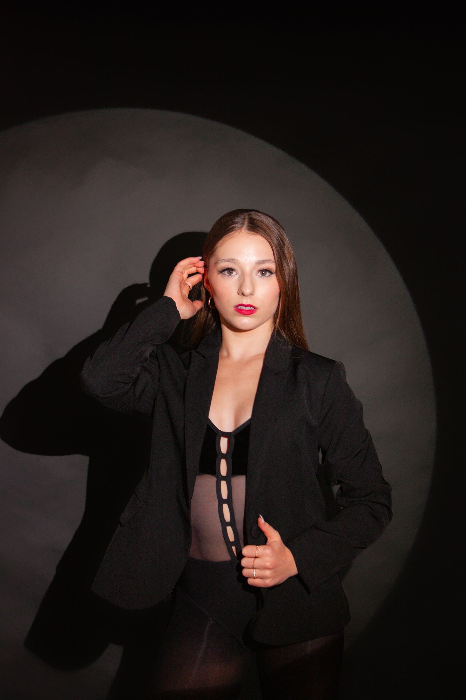Young woman in black blazer and bodysuit poses under spotlight against dark dramatic background.