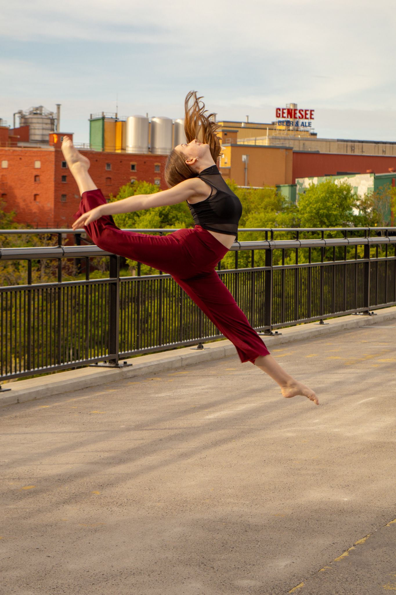 Dynamic dancer leaps mid-air near Genesee Brewery in black top and red pants outdoors.