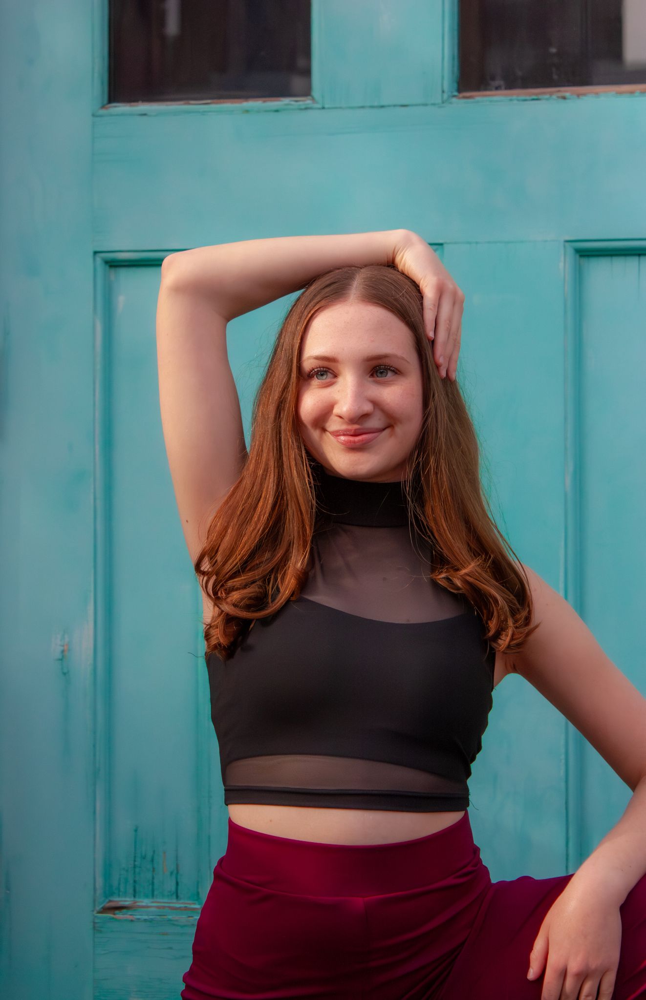 Smiling young dancer with auburn hair in black crop top poses against turquoise door backdrop.