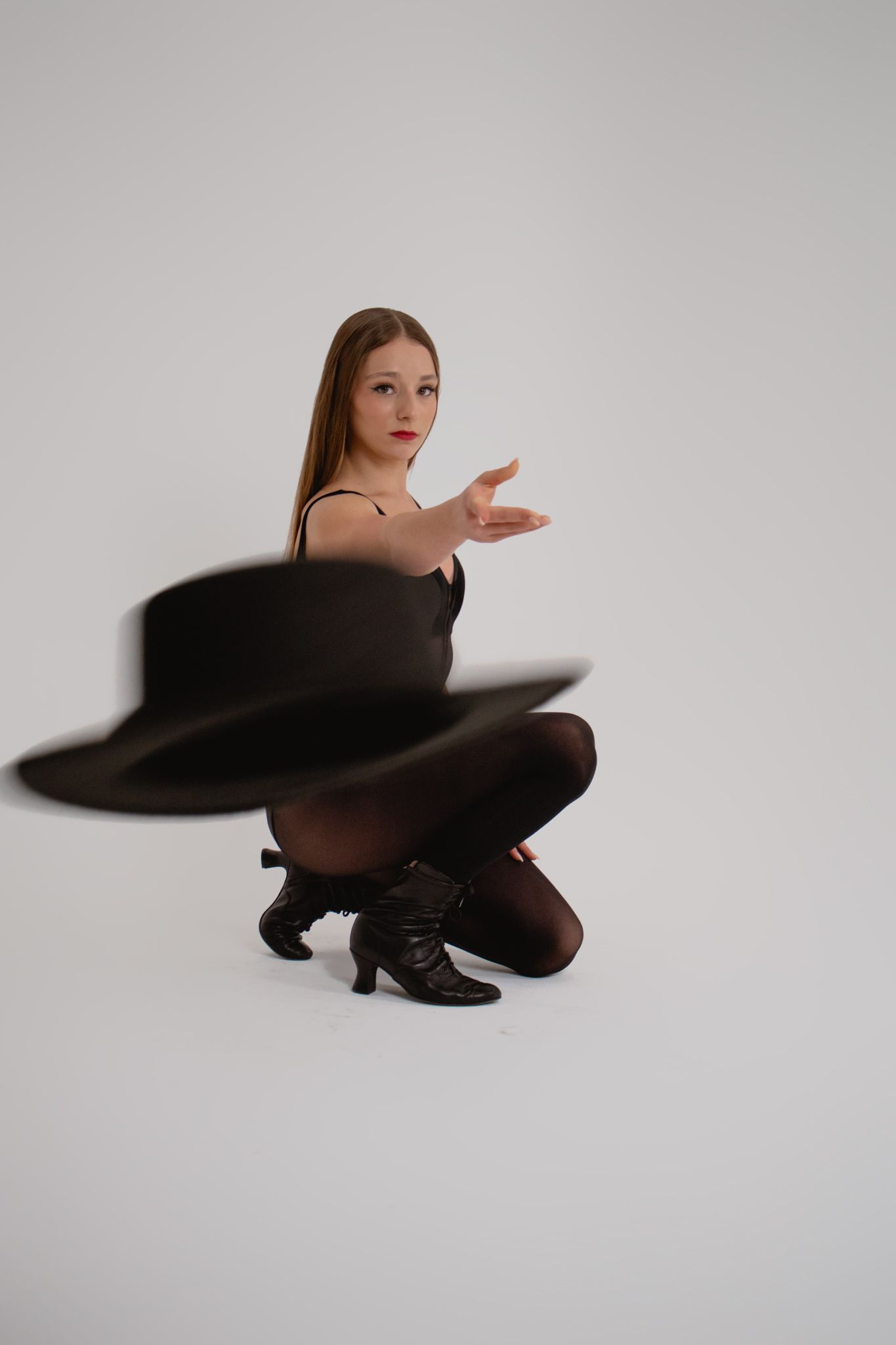 Female dancer in black outfit crouches while spinning a black hat in white studio setting.