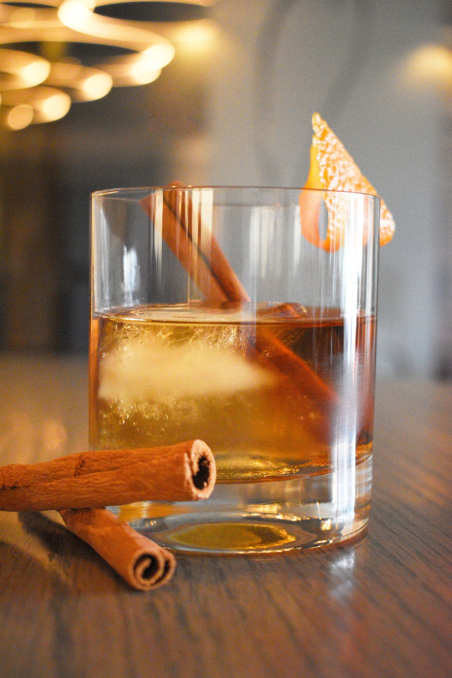 Whiskey cocktail with large ice cube garnished with cinnamon sticks and dried orange peel twist.