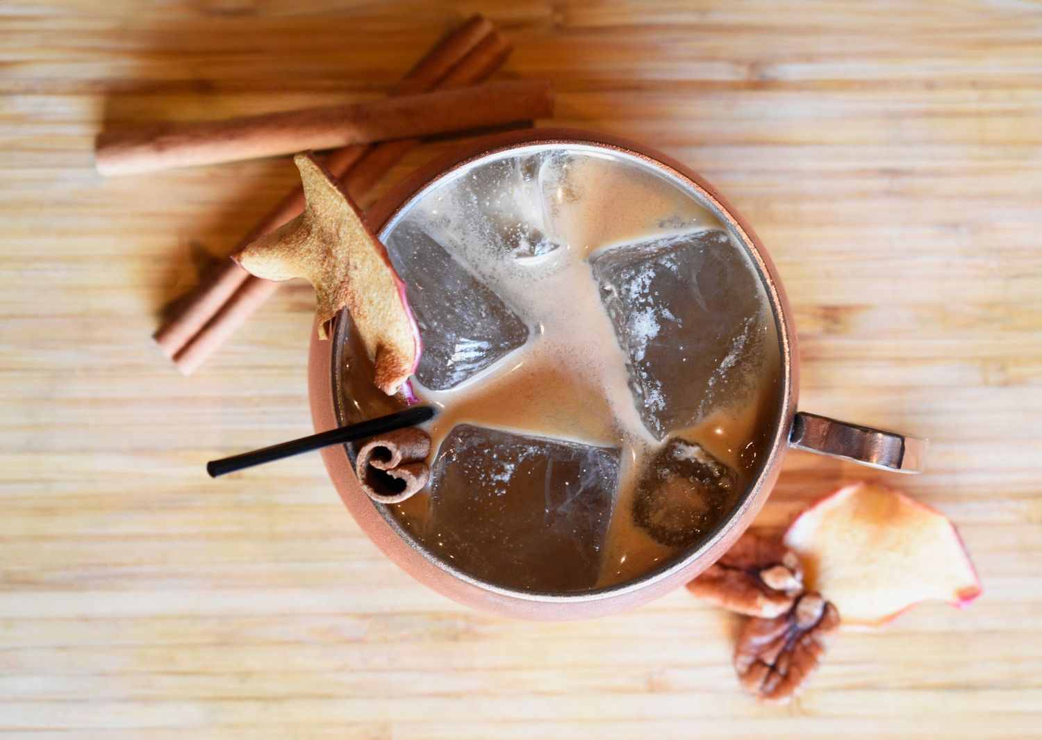 A copper Moscow mule mug with ice, cinnamon sticks, and dried fruit garnish on a wooden surface.
