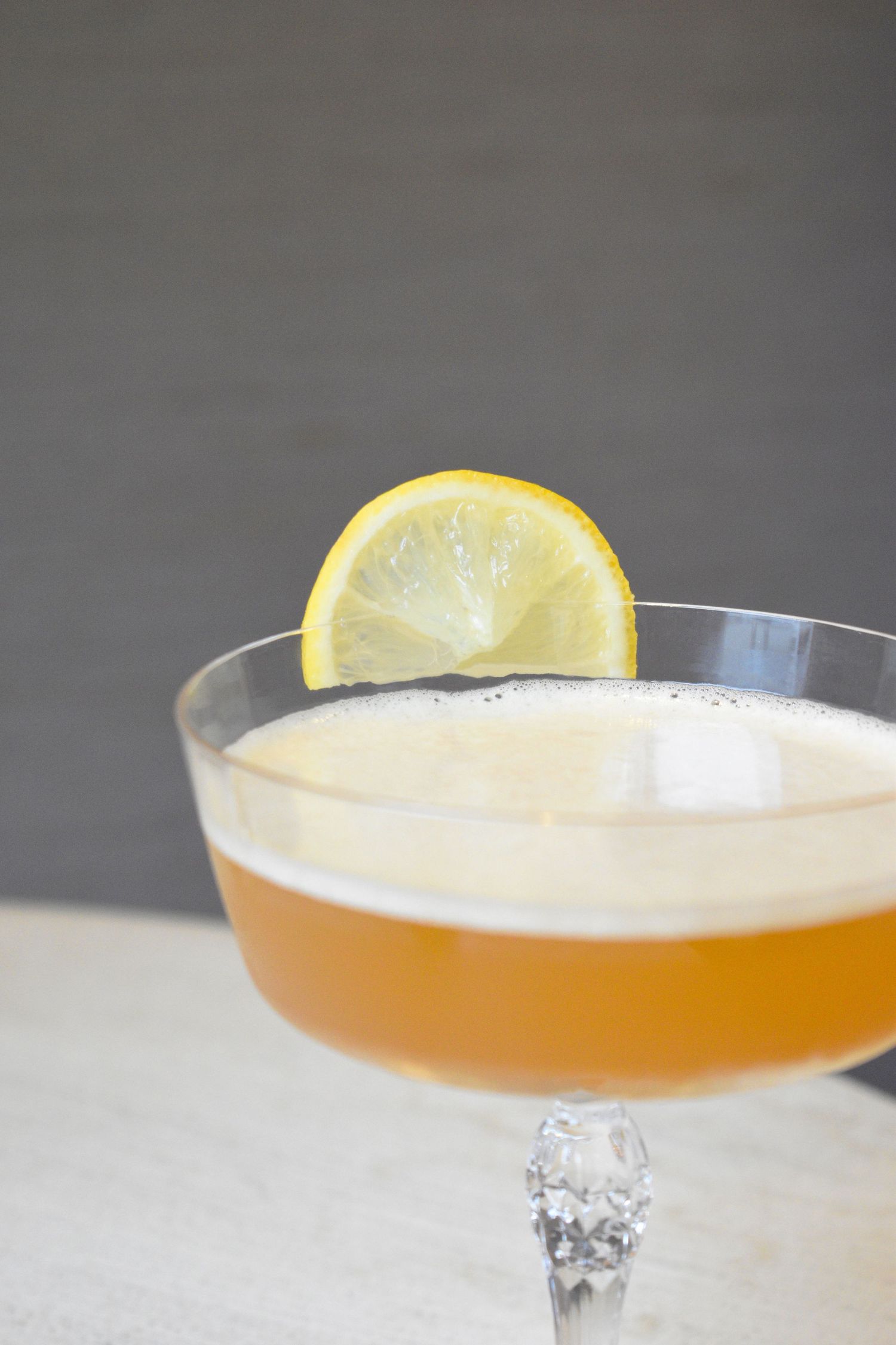 Close-up of a frothy amber cocktail in a crystal coupe glass topped with a fresh lemon wheel garnish.