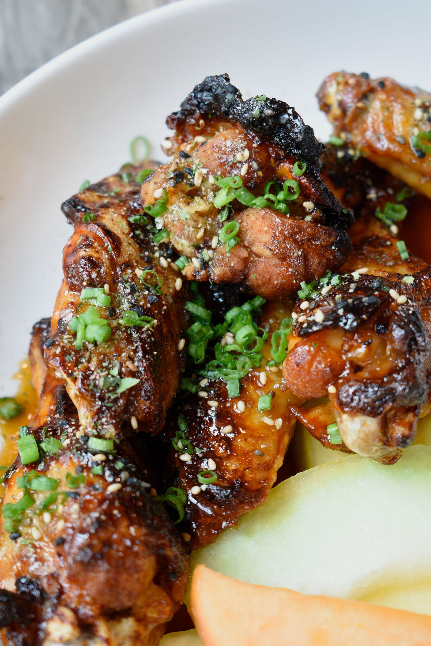 Glazed chicken wings topped with sesame seeds and green onions served with cucumber slices in a white bowl.