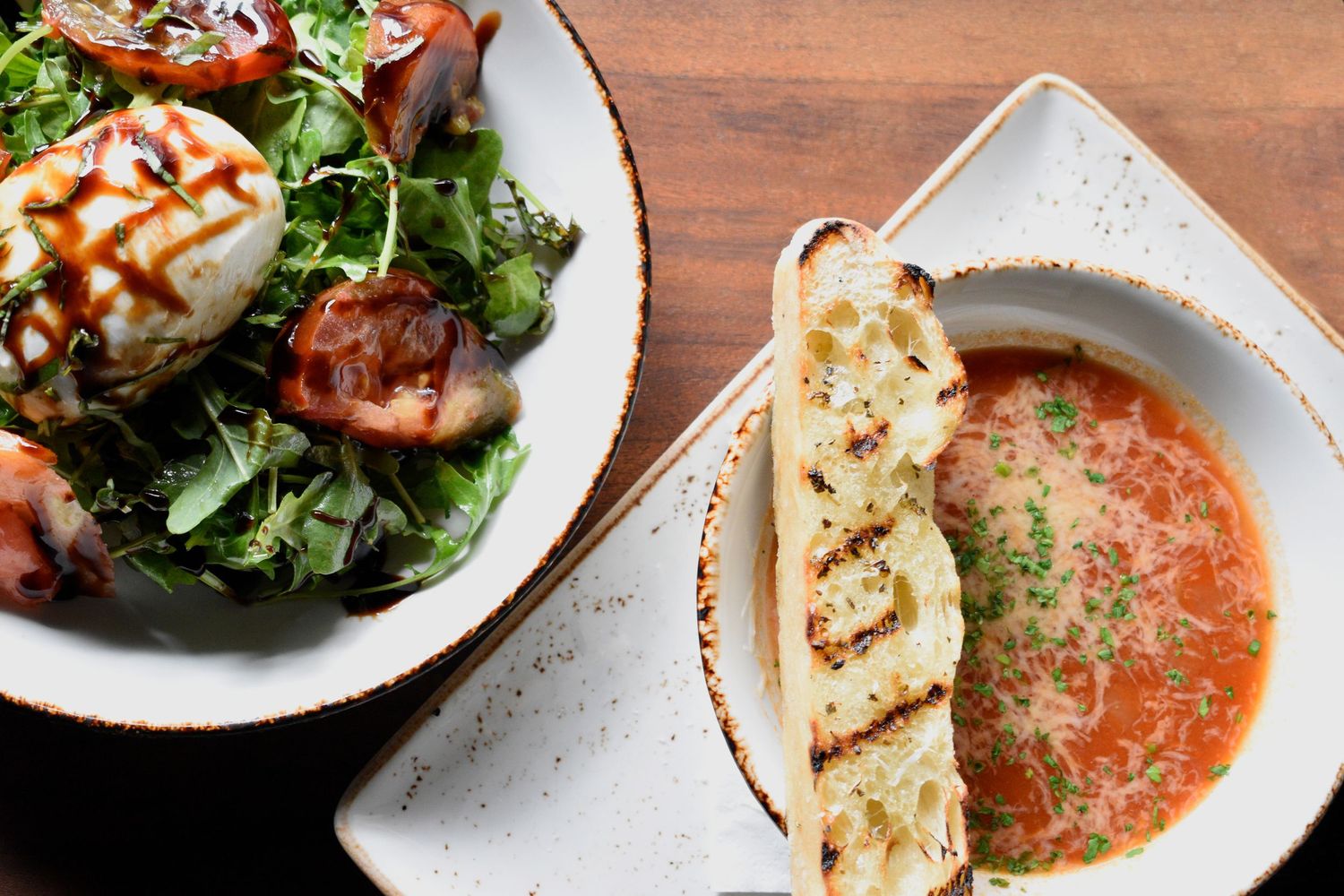 Tomato bisque soup with grilled flatbread alongside a burrata caprese salad on a wooden table.
