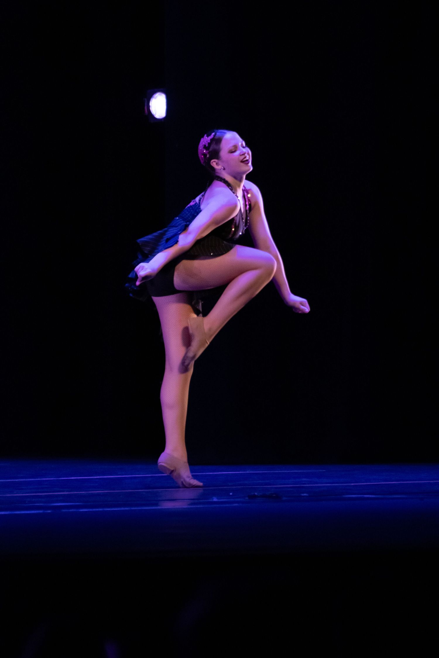 Expressive dancer in black costume performs high knee lift under spotlight on dark stage.