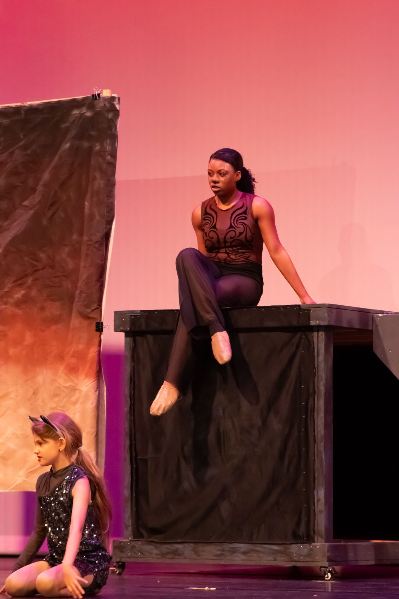 A dancer sits atop a prop box on stage while a young performer sits below in pink lighting.