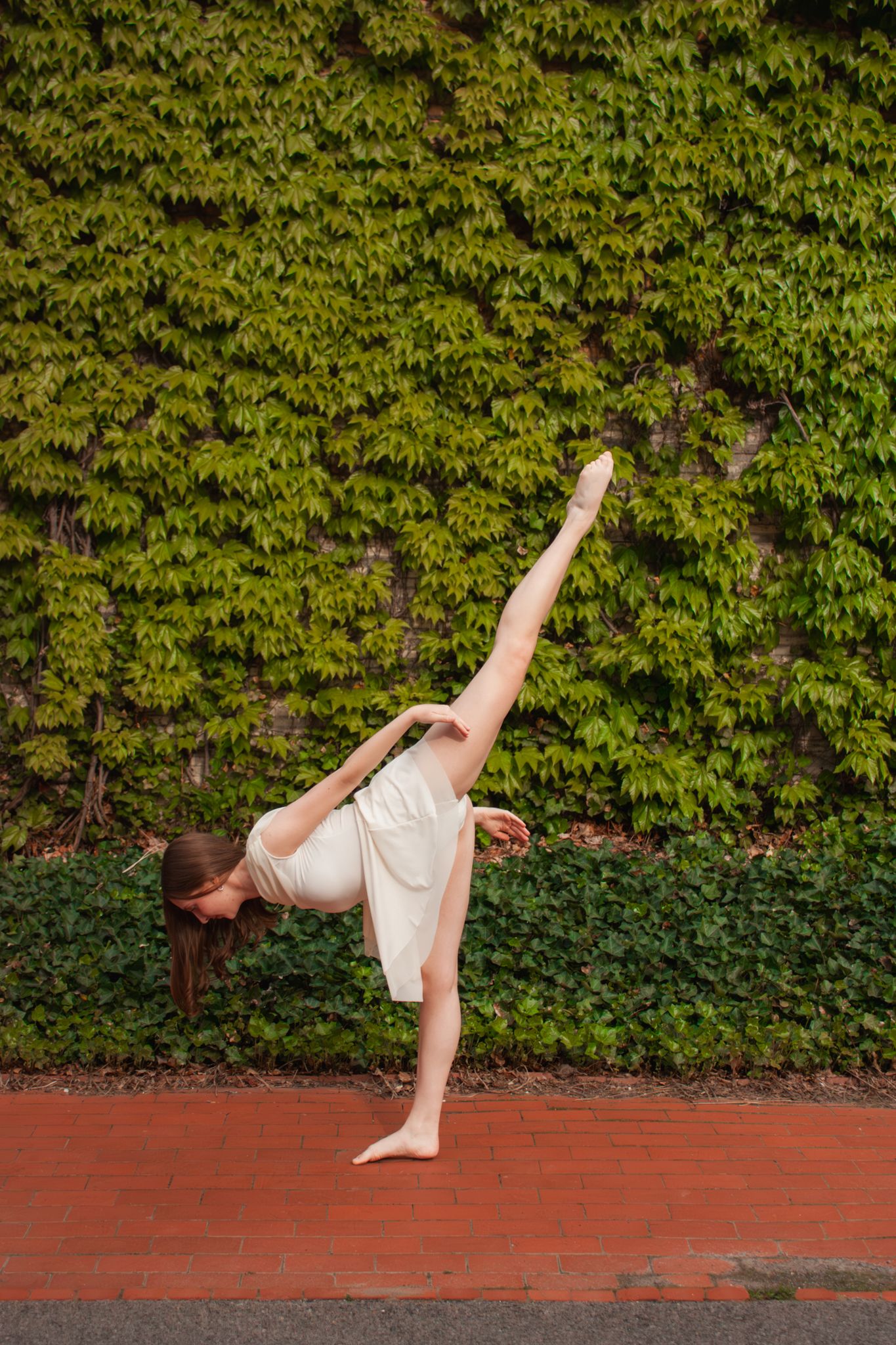 A flexible dancer executes a standing split pose barefoot on a red brick floor against an ivy wall.