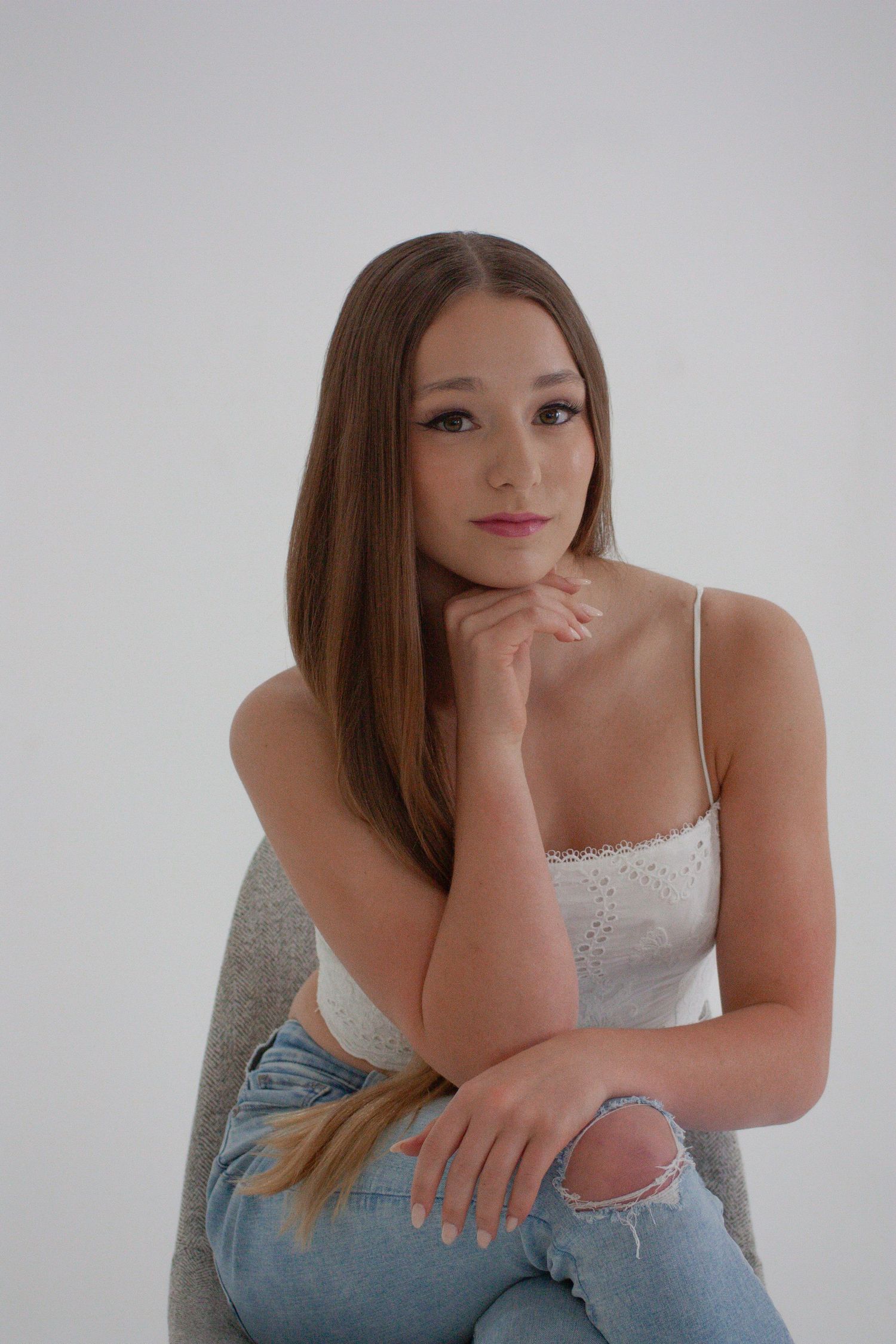 Young woman with straight brown hair sits pensively in white cami top and ripped jeans in studio.
