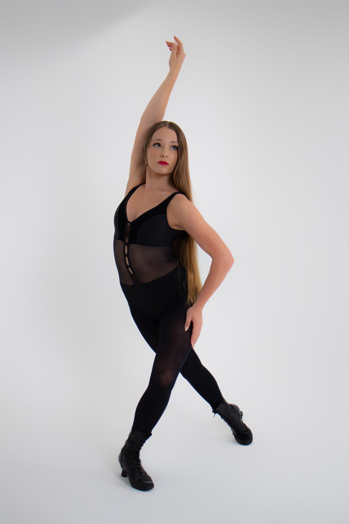 Female dancer in black bodysuit and tights performs ballet pose with raised arm in studio.