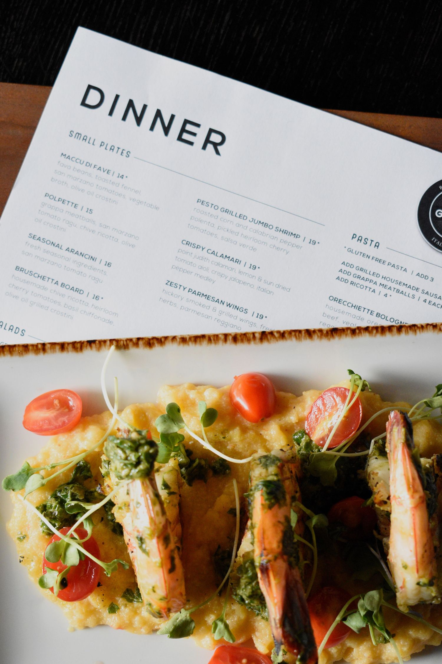 Pesto grilled jumbo shrimp over creamy polenta with cherry tomatoes beside a dinner menu.