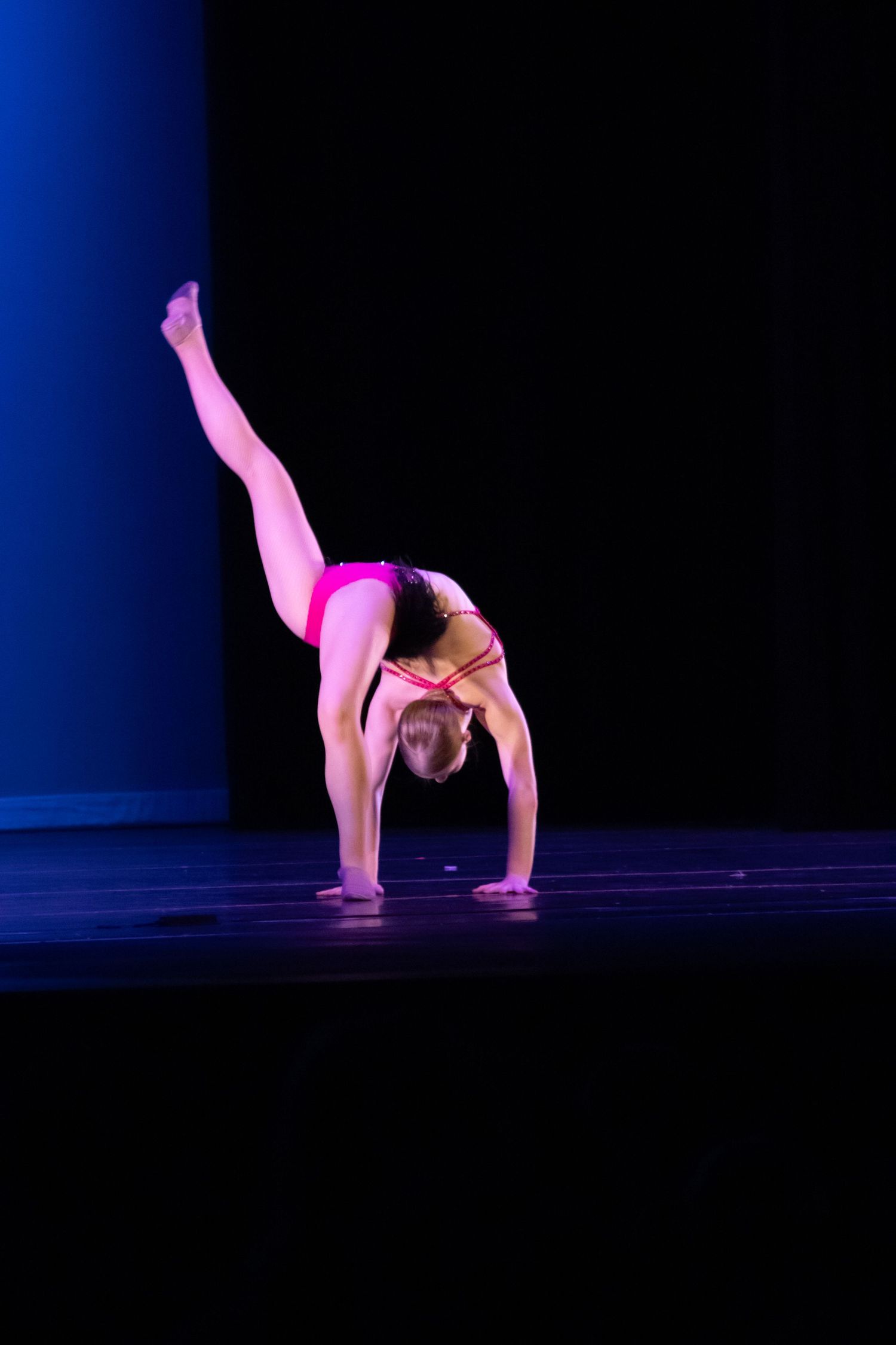 Flexible dancer in pink leotard performs a back walkover with leg raised on dark stage.