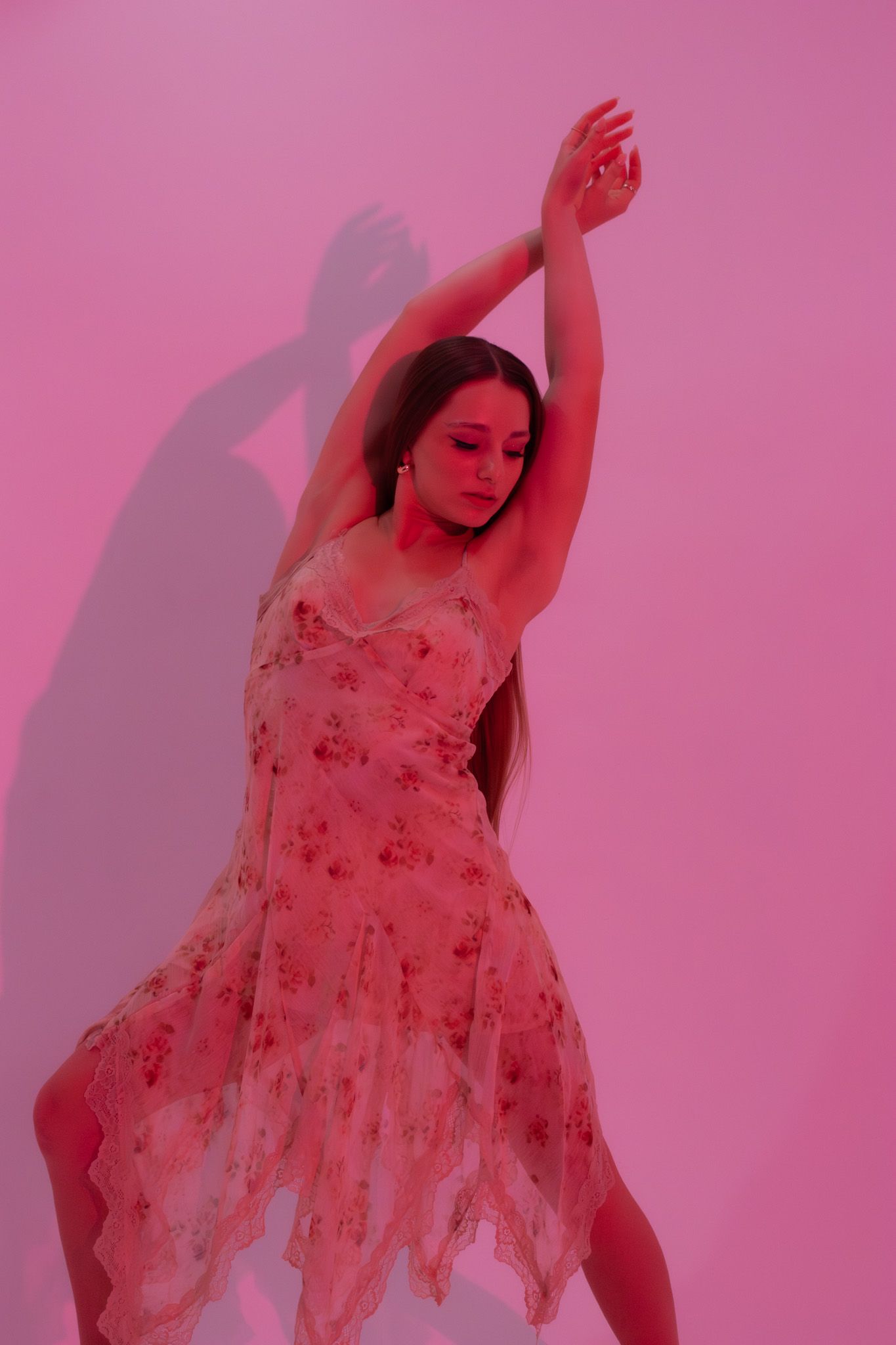 Young woman in floral dress dances with arms raised against vibrant pink studio background.