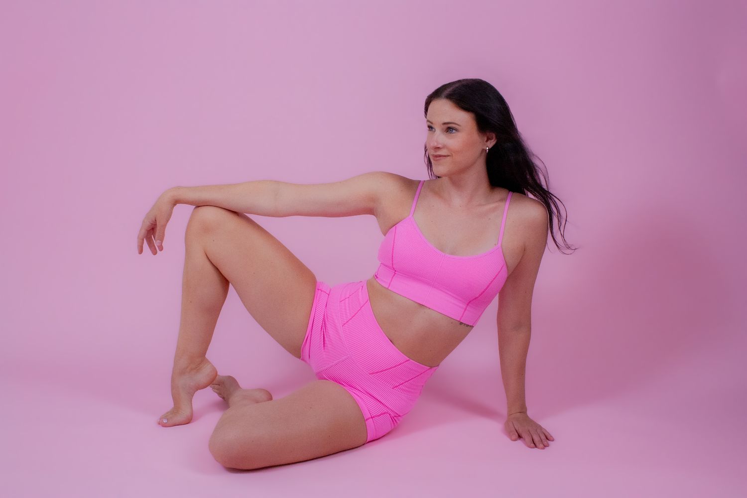 Woman in pink crop top and shorts sitting on pink studio floor.