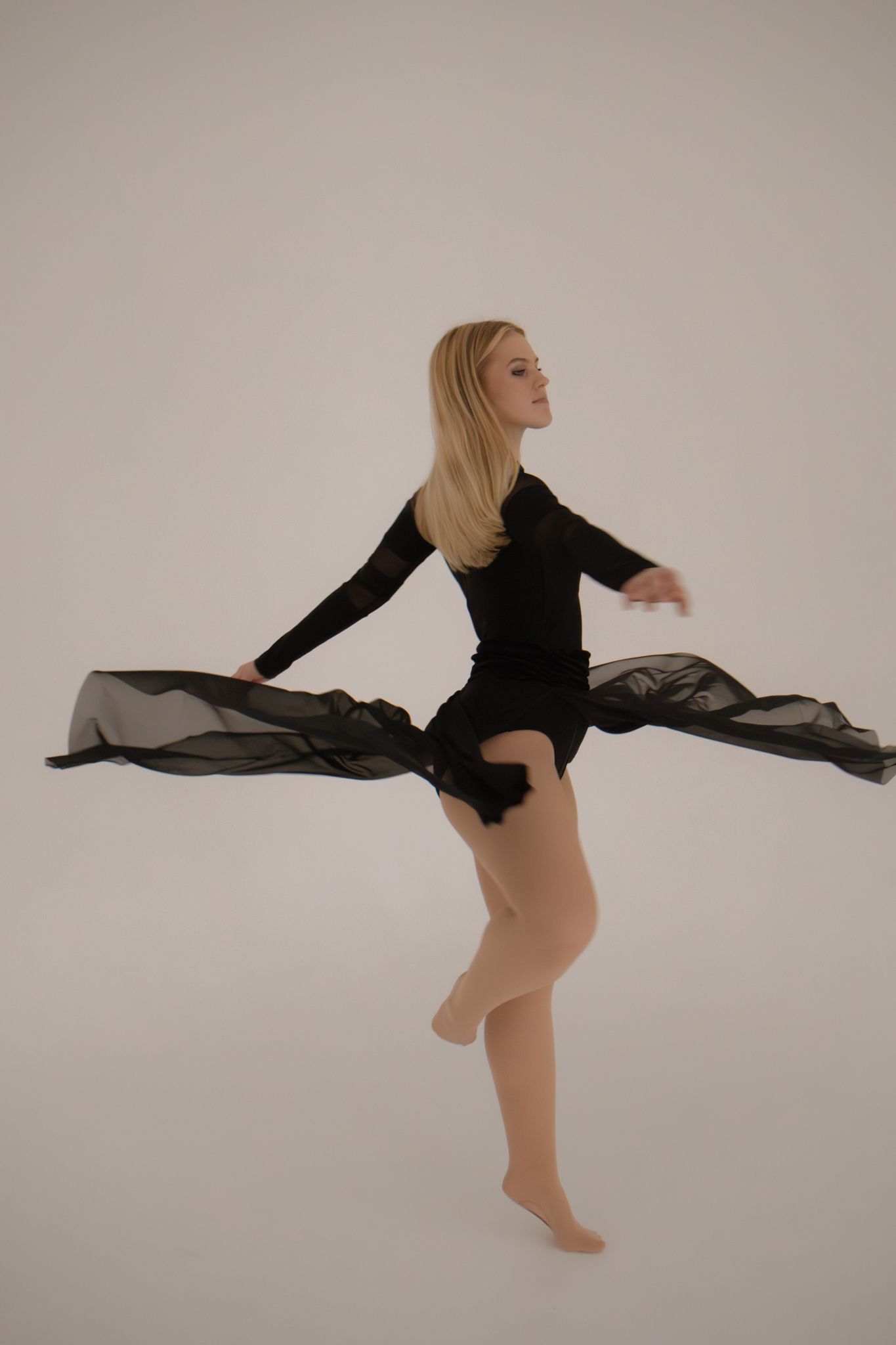 Blonde dancer in black bodysuit with flowing sheer fabric captured mid-movement on white background.