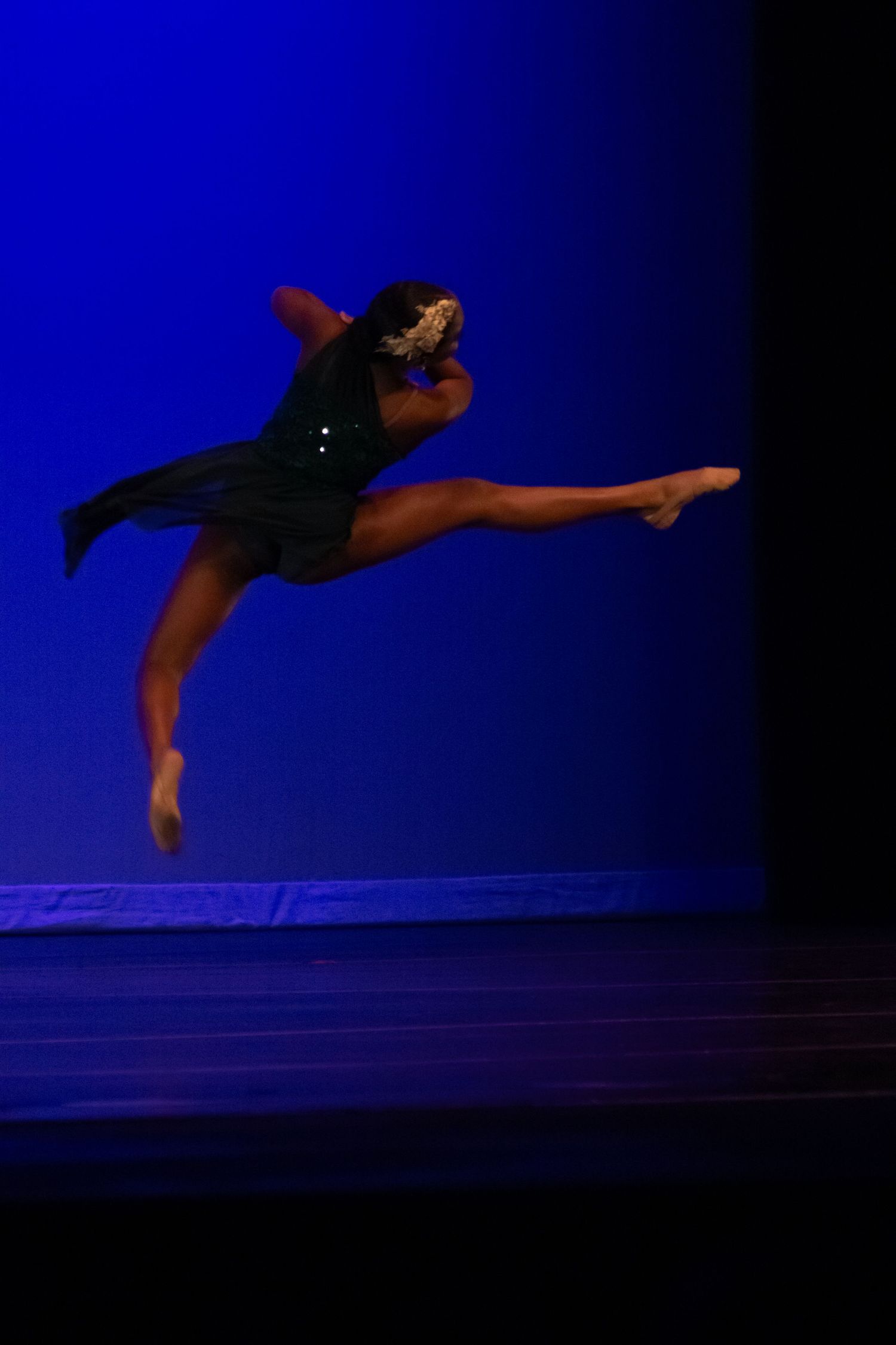 A dancer leaps gracefully in a dark green costume against a vivid blue stage backdrop.
