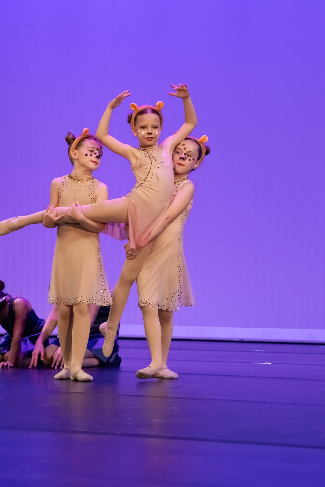 Three young girls in beige costumes perform a ballet routine on a purple-lit recital stage.