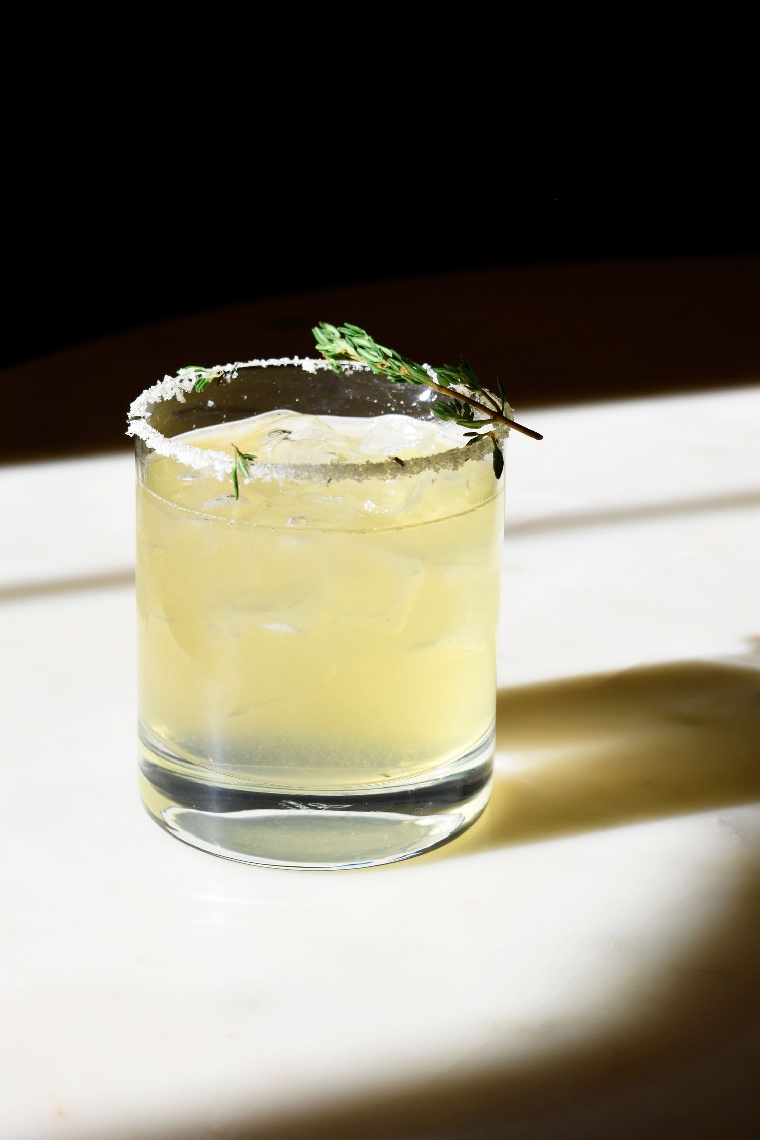 Refreshing pale yellow margarita with salted rim and fresh thyme garnish served in a rocks glass.