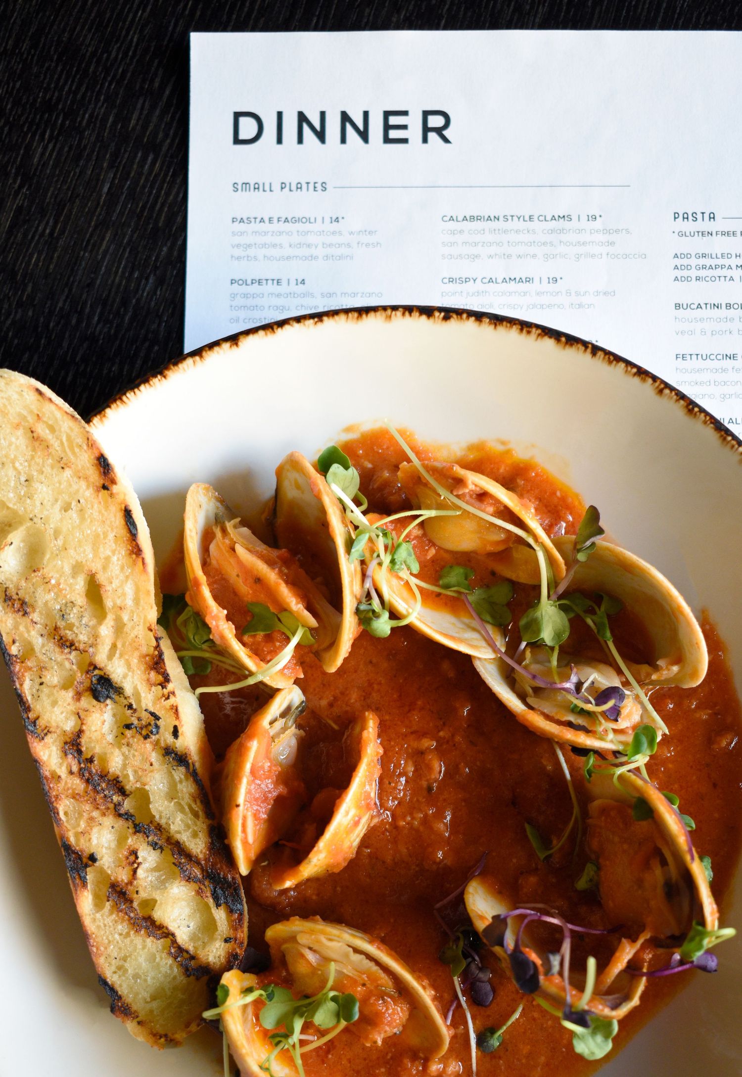 Calabrian-style clams in tomato broth with grilled bread served beside a dinner menu card.