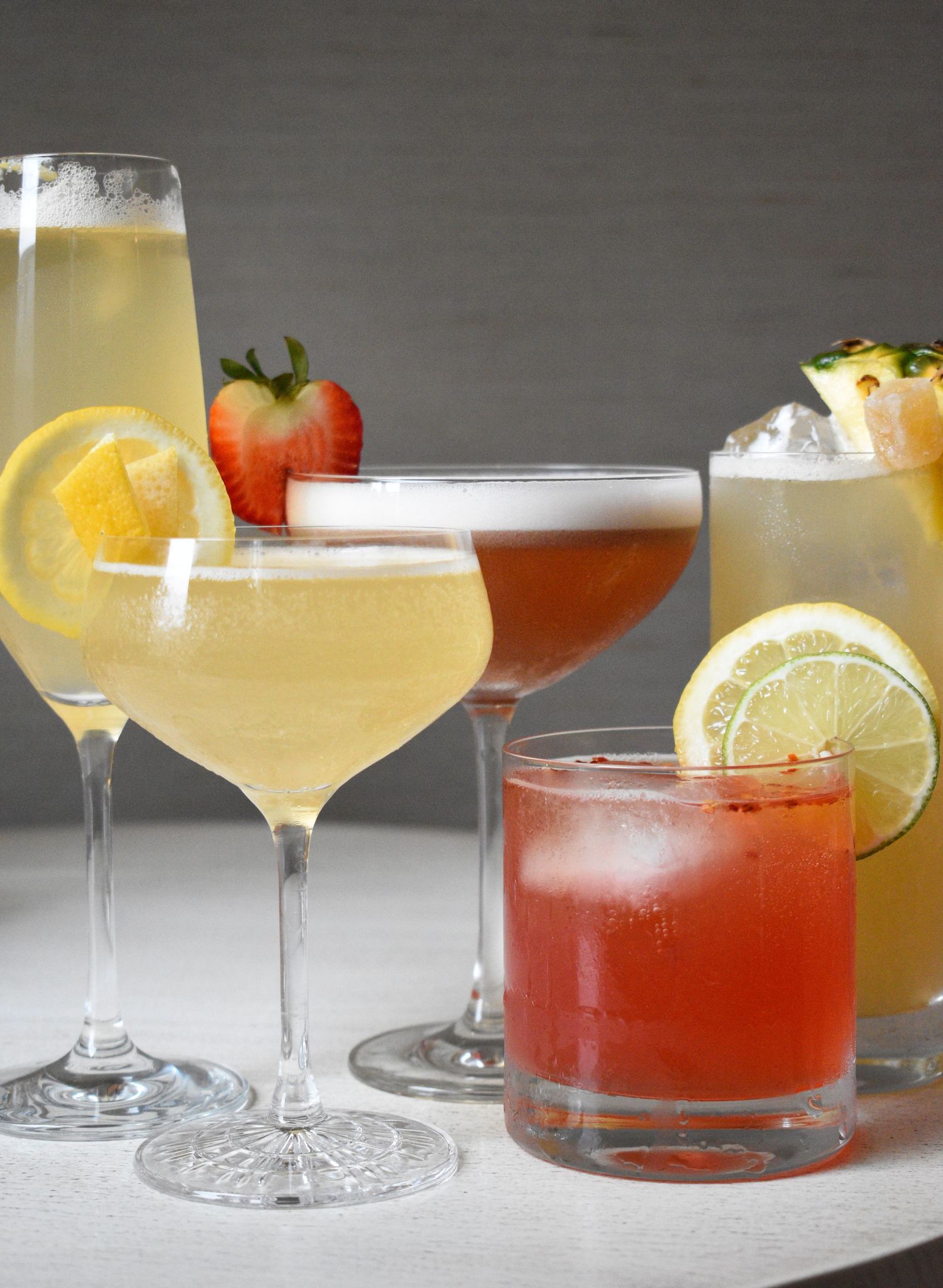 Assorted cocktails including champagne flute, coupe, and rocks glasses garnished with fresh fruit on white surface.