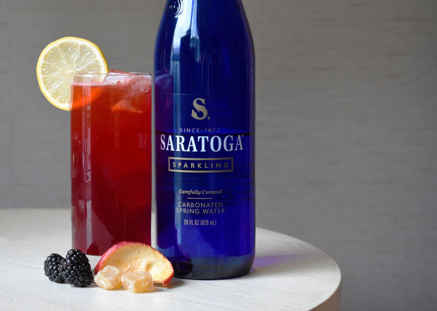 Vibrant red cocktail with lemon wheel garnish next to a blue Saratoga sparkling water bottle on a white surface.