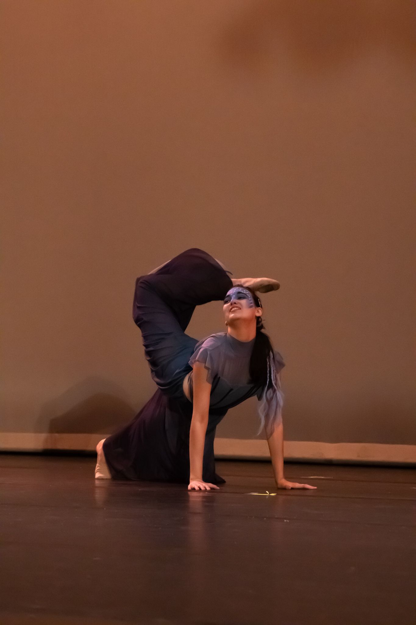 A solo dancer in dark clothing performs an advanced backbend floor pose during a contemporary dance recital.
