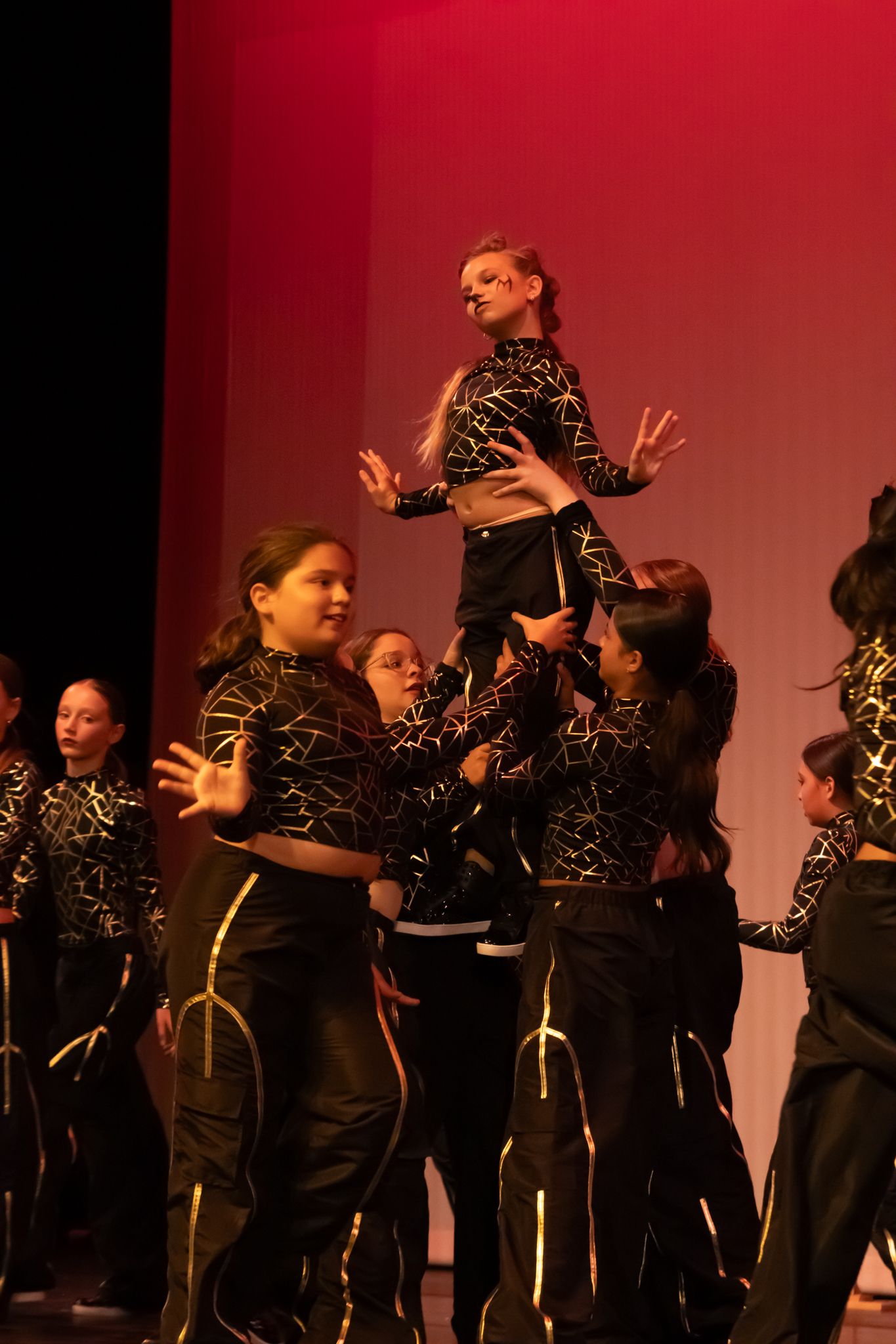 Group of dancers in black and gold costumes lift a performer during an energetic stage recital.