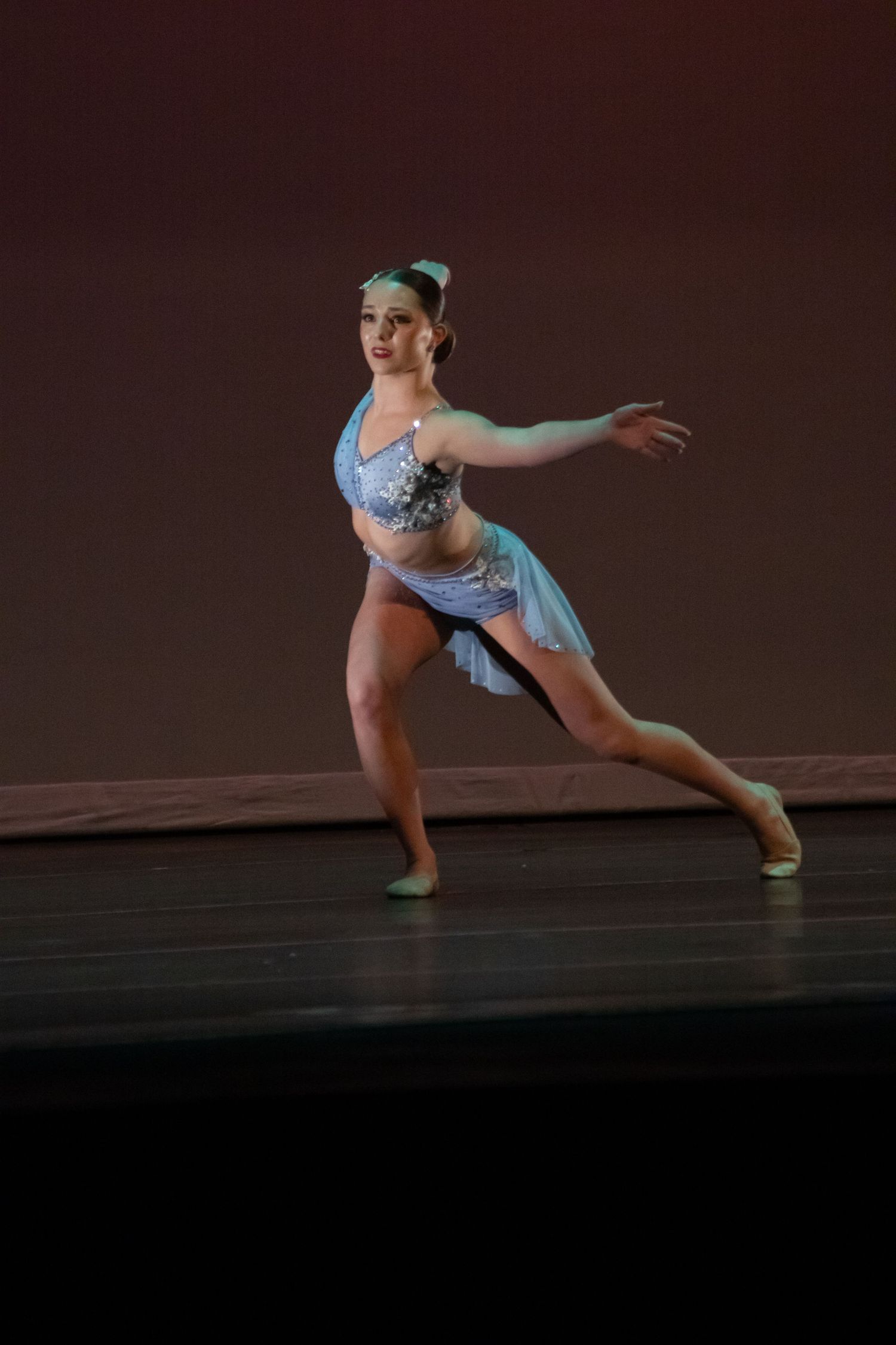 A young dancer in a light blue costume performs an elegant lunge pose on a warmly lit stage.