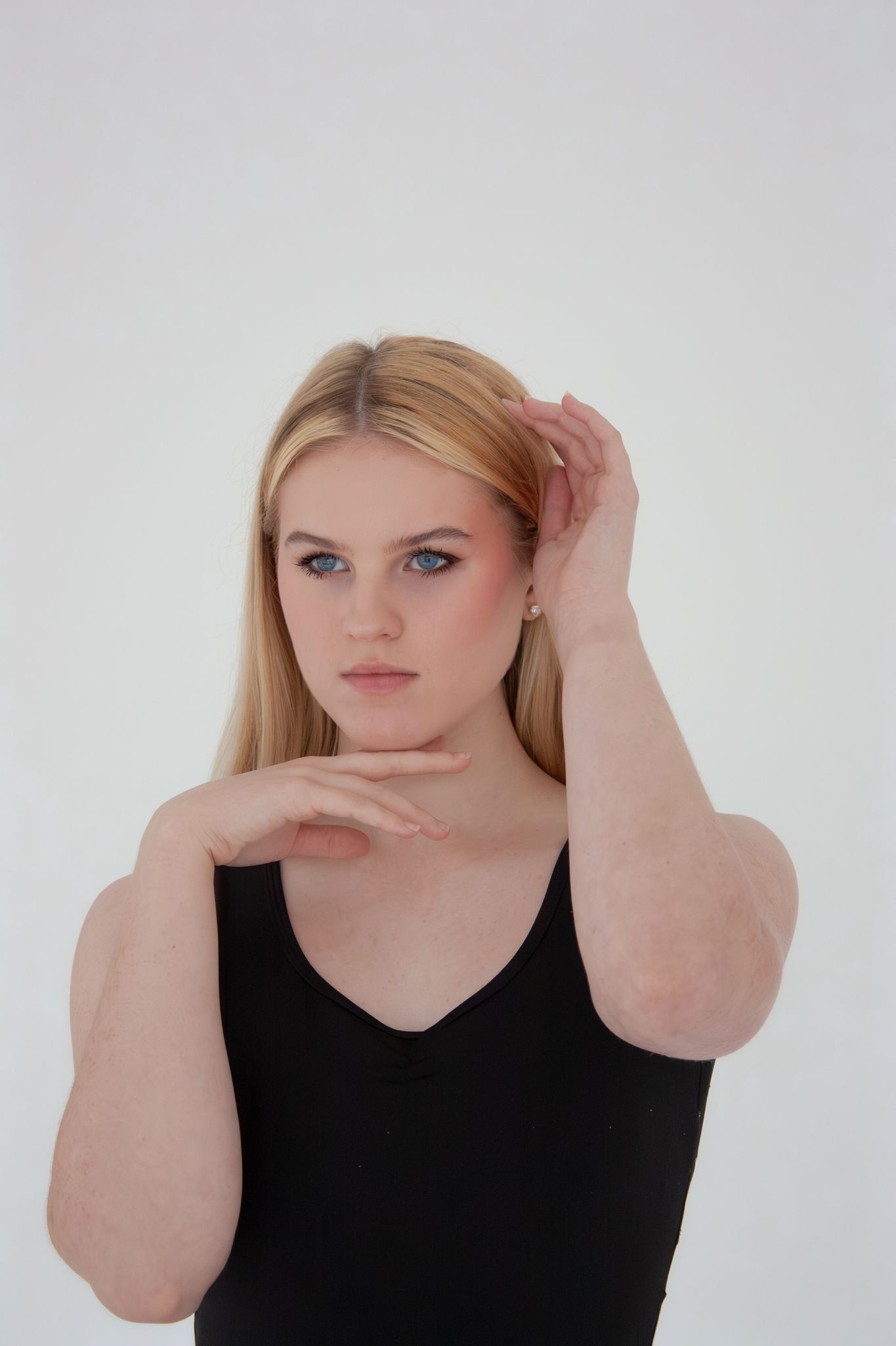 Blonde blue-eyed woman in black top poses with hands raised near face against white background.
