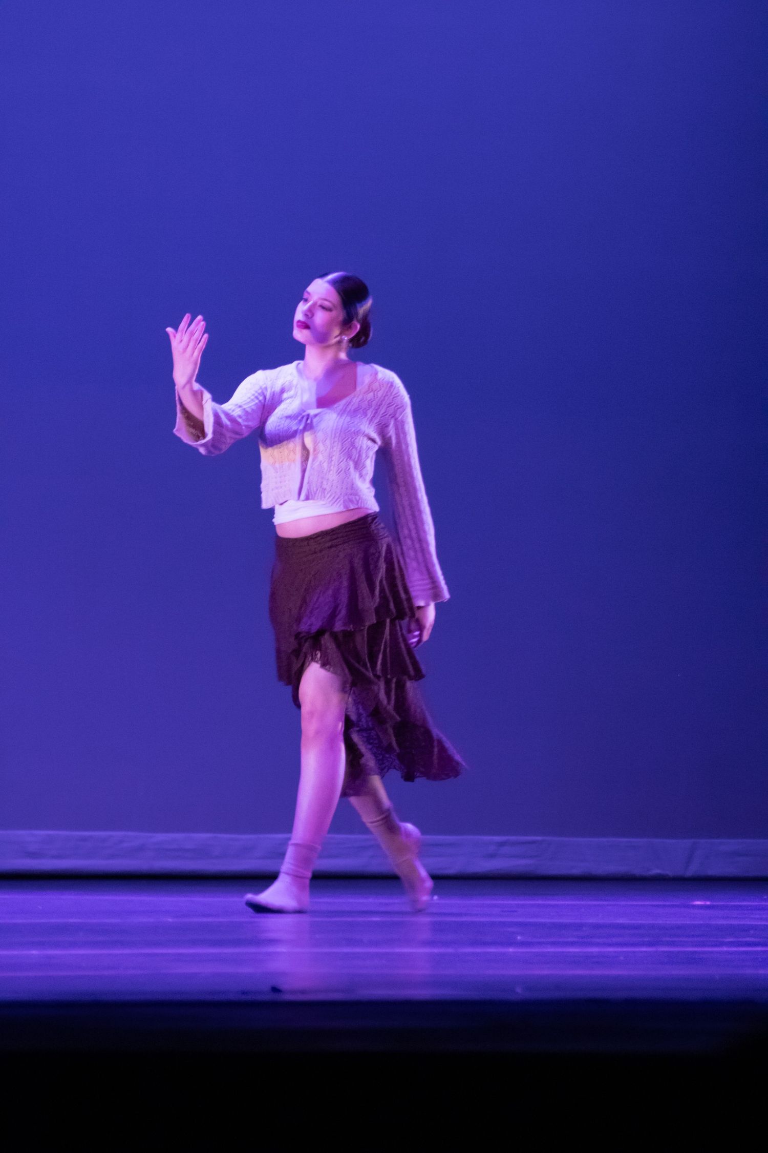 A female dancer performs a contemporary solo on stage in a white top and brown skirt under blue lighting.